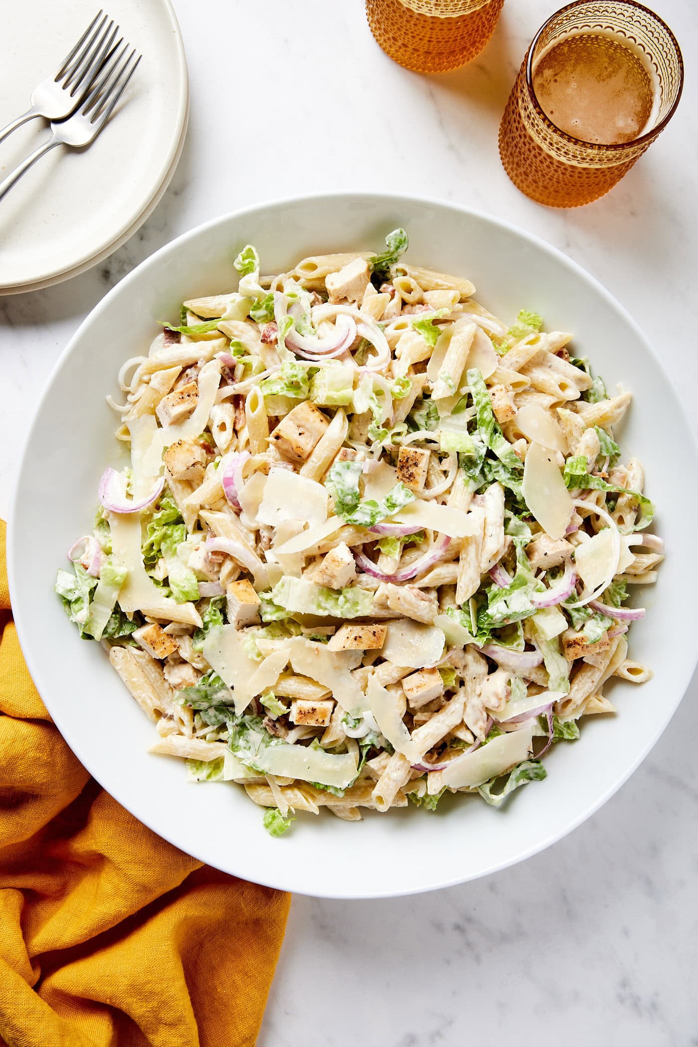 Large white bowl on white marble surface filled with chicken Caesar pasta salad with two glasses filled with liquid to the side.