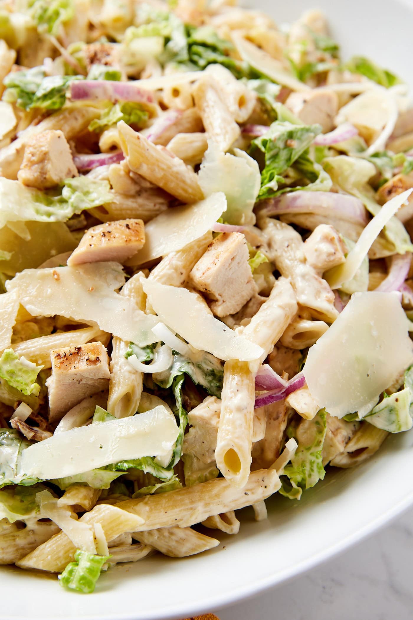 Large white bowl filled with chicken Caesar pasta salad with shards of Parmesan cheese, pasta, red onion, lettuce, and more.