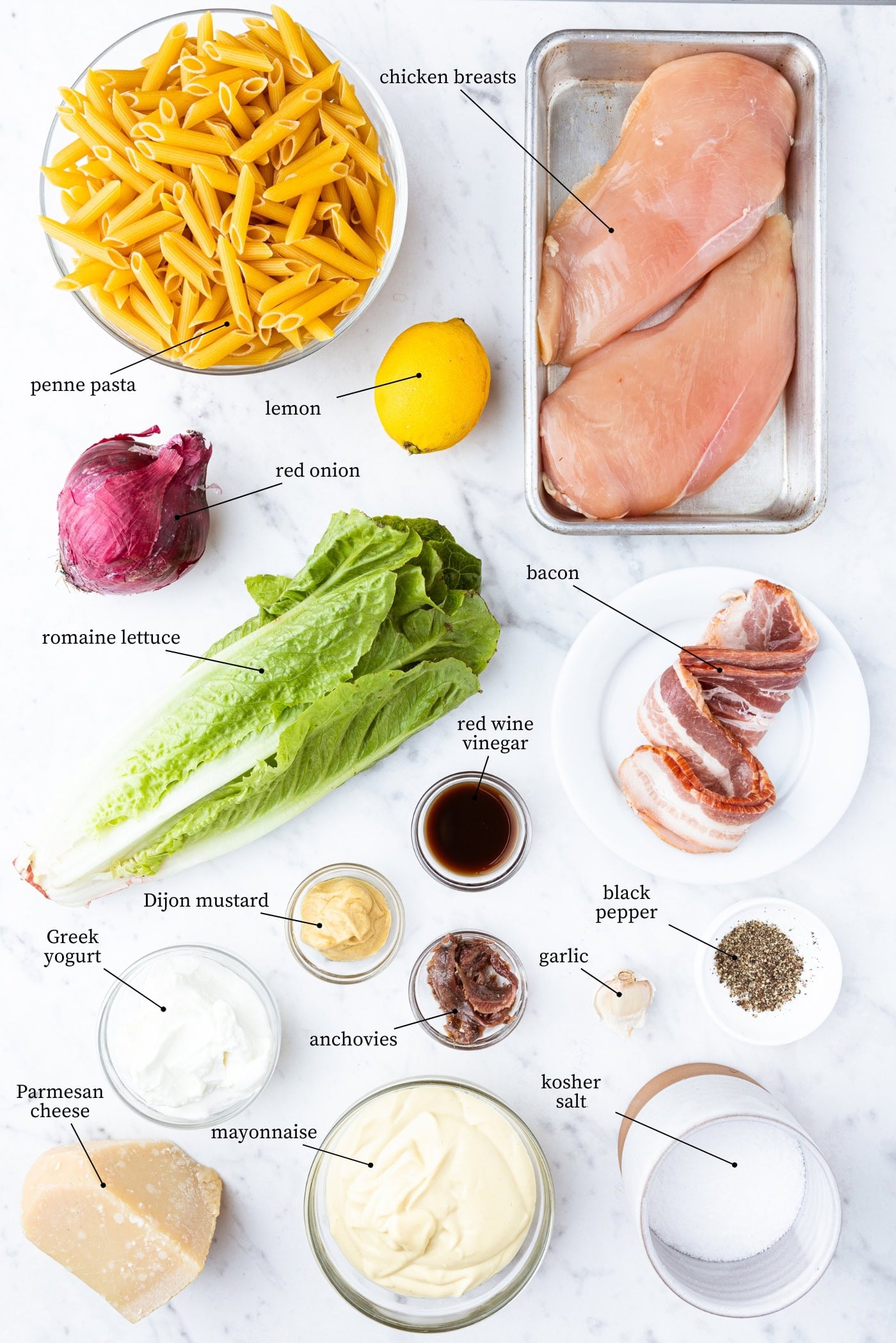 White marble surface filled with all ingredients needed for a chicken Caesar pasta salad including pasta, lettuce, bacon, chicken, and more.