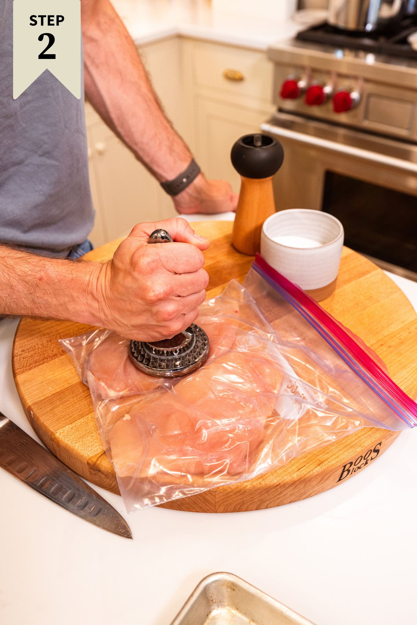 Hand holding heavy mallet pounding raw chicken breasts in a plastic bag to flatten them.
