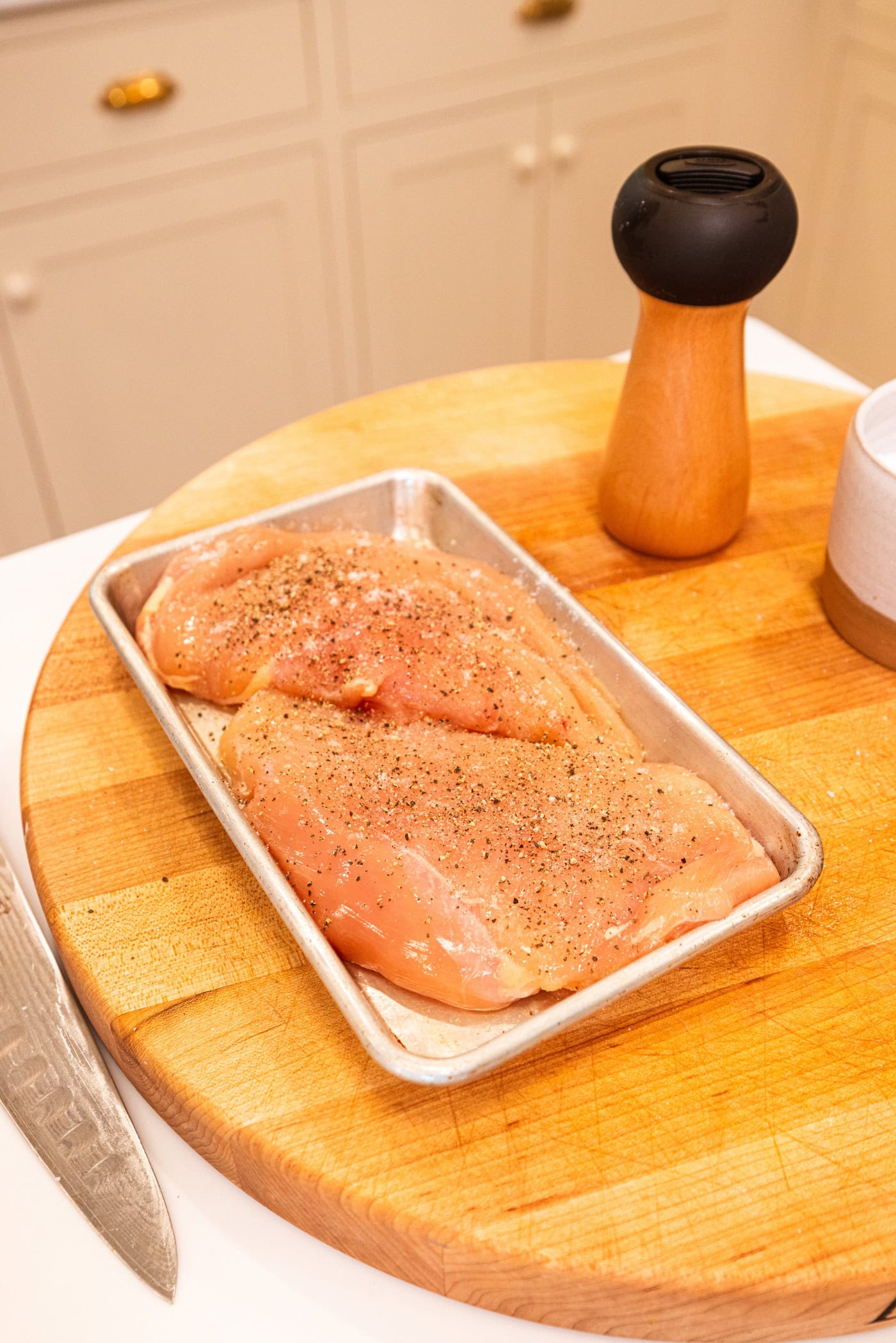Two raw chicken breasts with salt and black pepper on top sitting on a baking pan before being cooked.