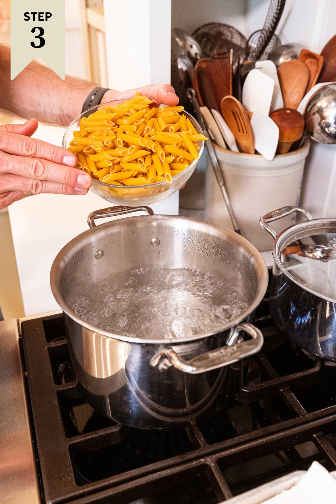 Hand holding glass bowl filled with uncooked penne pasta pouring into a pot with boiling water.