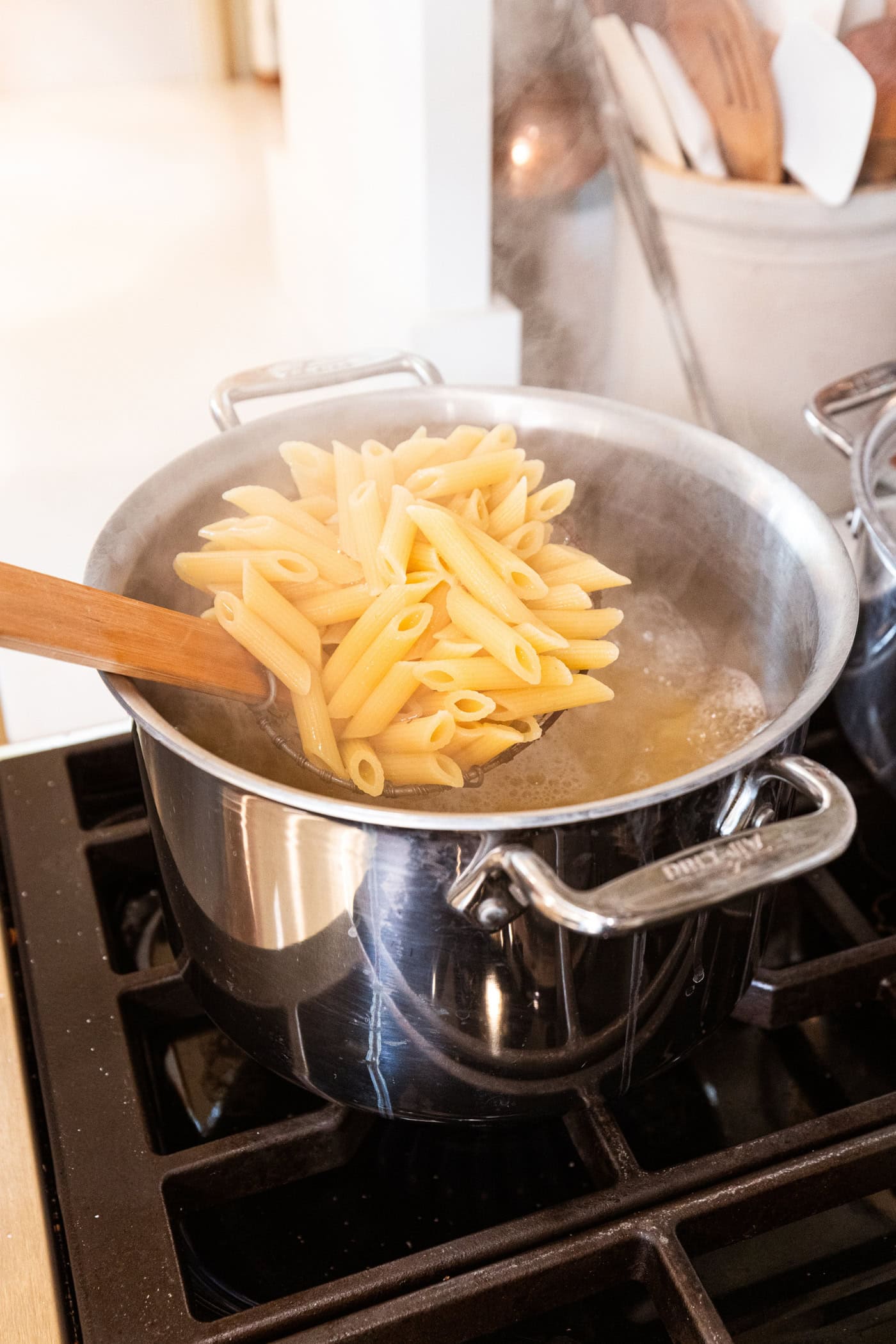 Ladle holding up cooked penne pasta from a large stockpot on the stove.