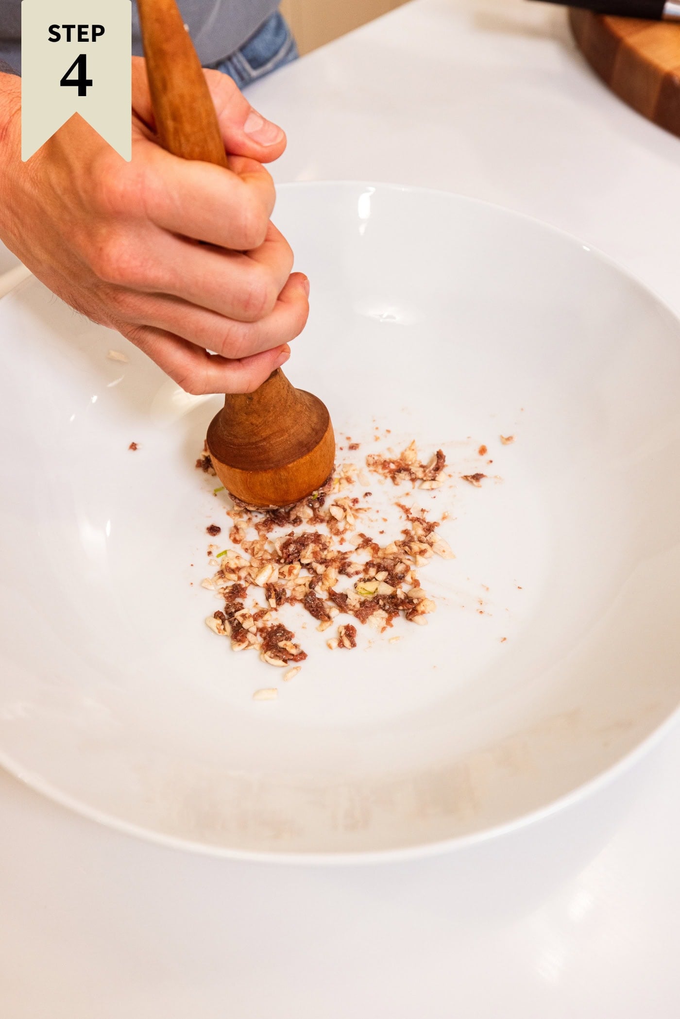 Large white bowl with a wooden mallet smashing pieces of anchovy and garlic to make a Caesar dressing.
