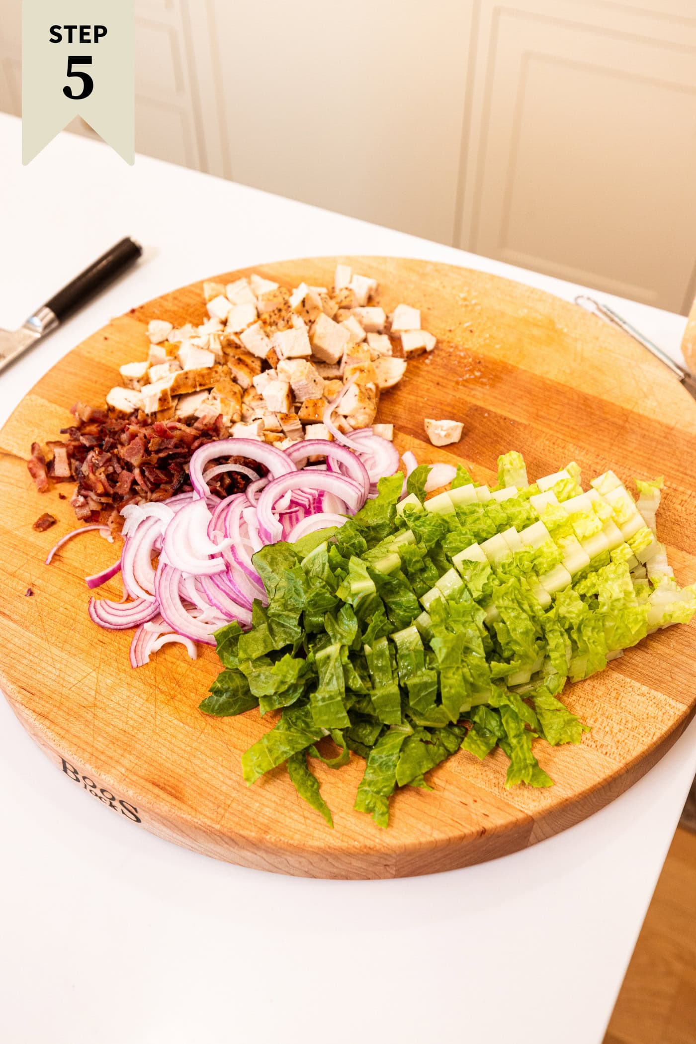 Toppings for a chicken Caesar pasta salad including red onion, lettuce, bacon, and cubed chicken breast.