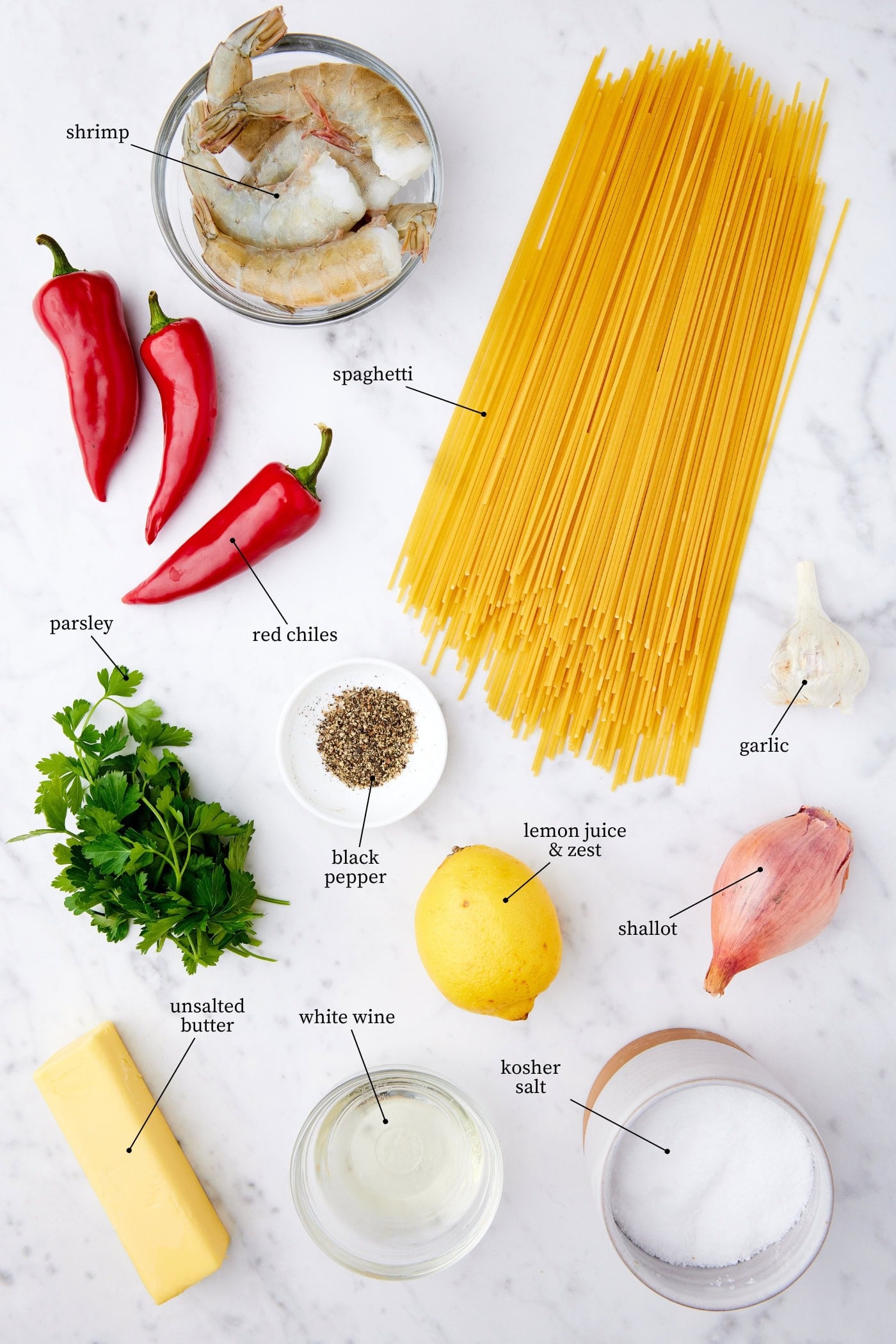 White marble surface filled with all ingredients needed for a garlic shrimp pasta recipe with spaghetti, chiles, raw shrimp, and more.