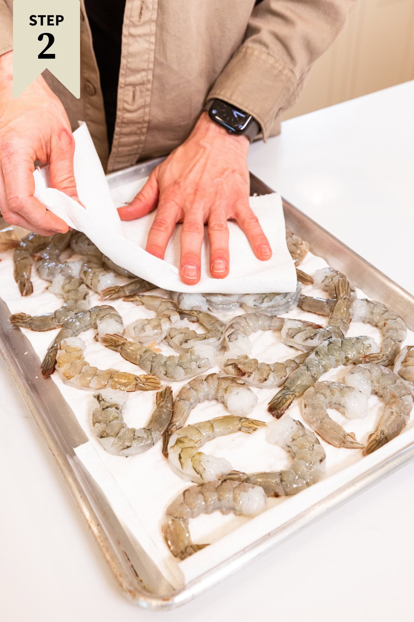 Hand using a paper towel to pat dry raw shrimp on a parchment-lined baking sheet.