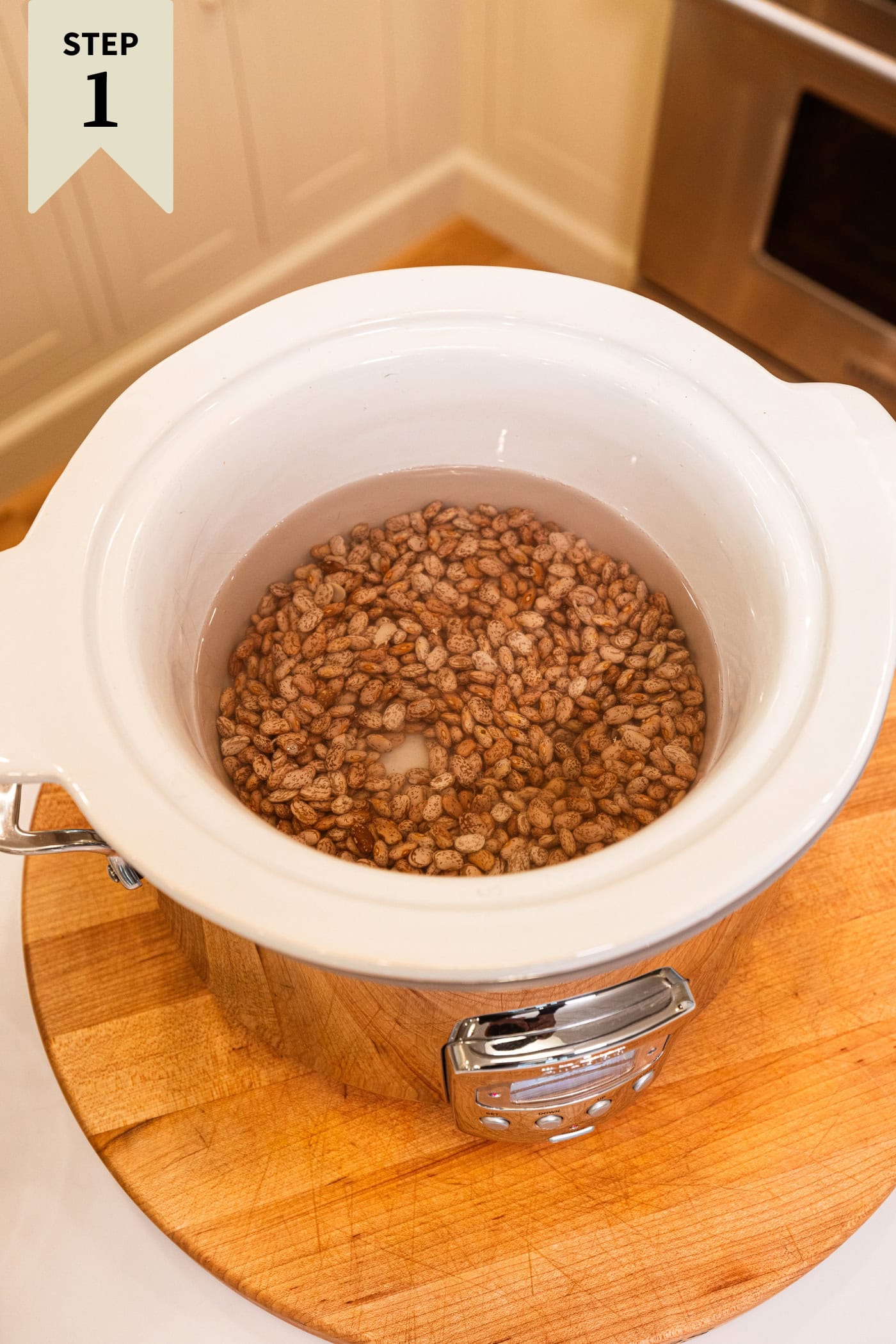Slow cooker with dried beans soaking in water on a wooden cutting board.