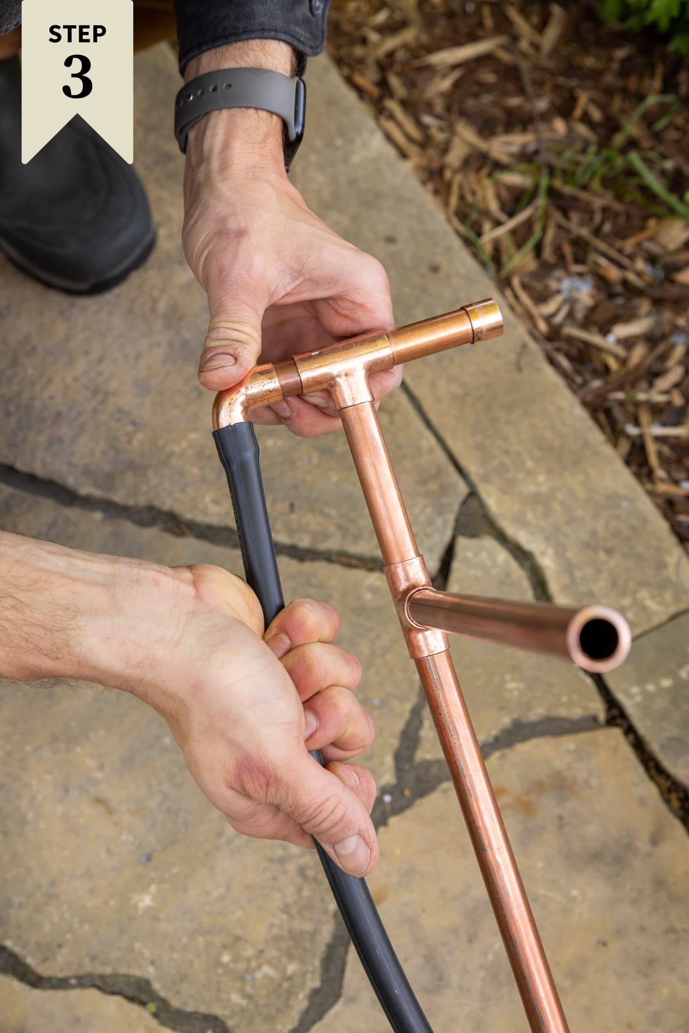 Hand placing a rubber hose into the end of a copper pipe for a DIY water fountain.