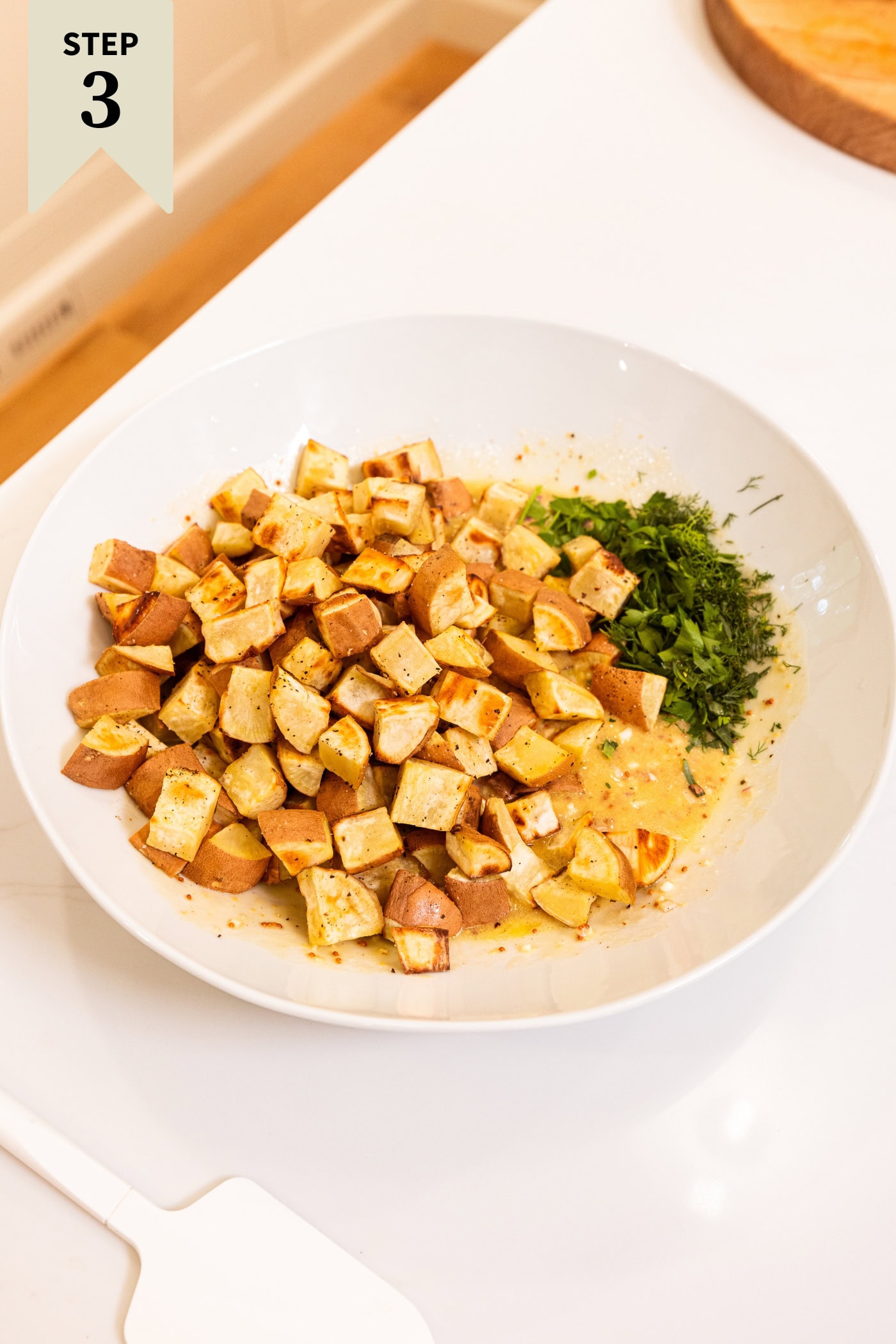 Large white serving bowl with herbs, vinaigrette, and roasted diced sweet potatoes before being mixed together.