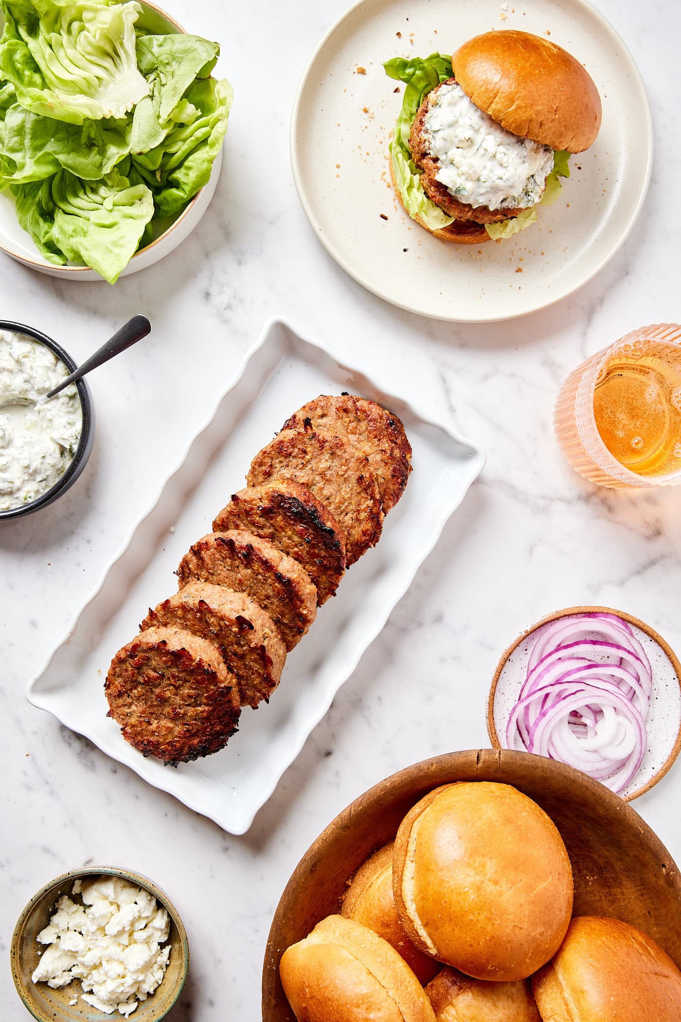 White marble surface with all ingredients needed to assemble turkey burgers including grilled burgers, buns, lettuce, tzatziki sauce, and more.