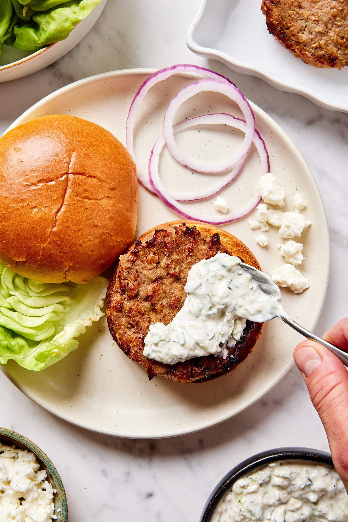 White plate with burger being assembled with homemade tzatziki sauce being spooned on top of burger with bun and lettuce and onions to the side.