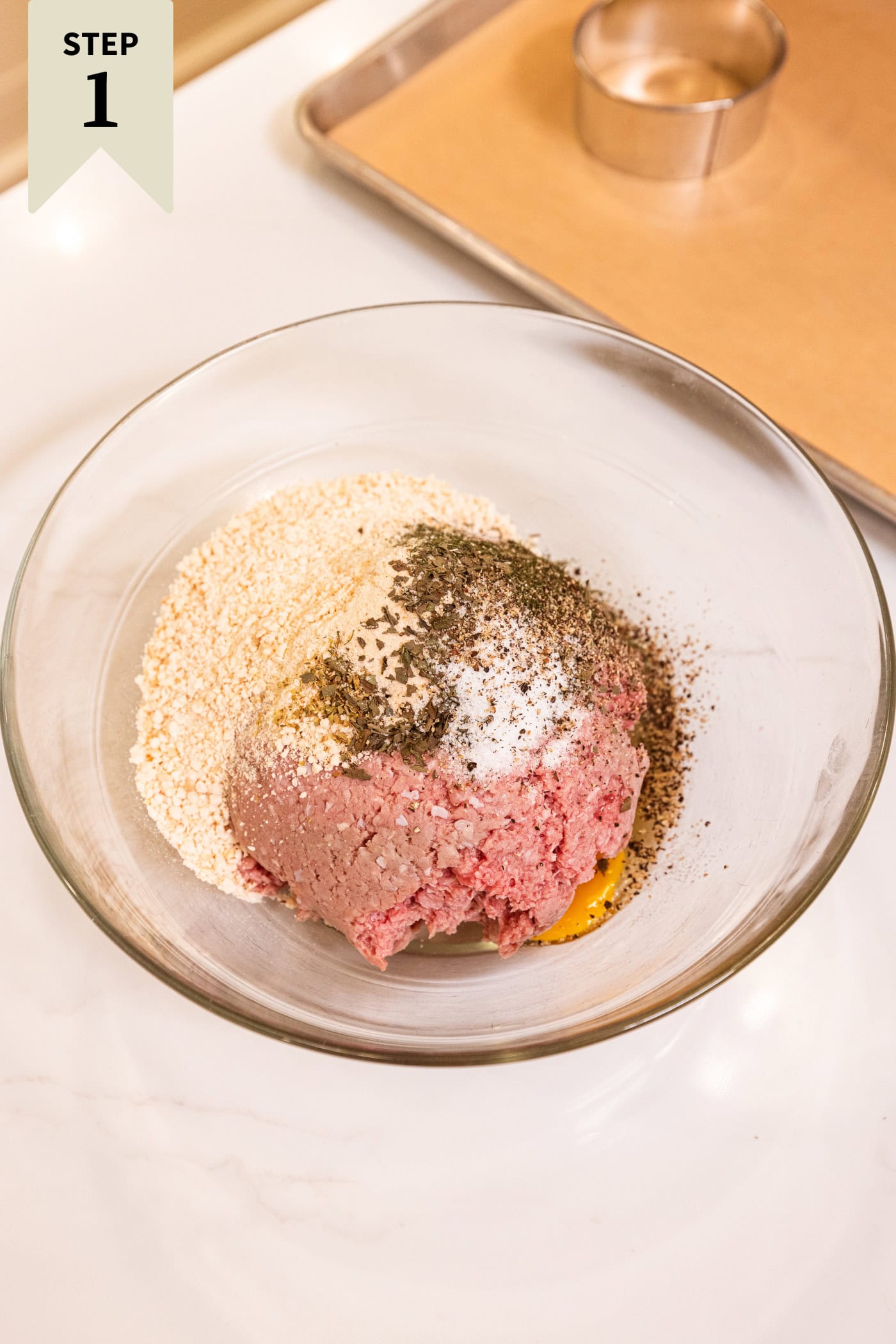 Glass bowl filled with ingredients needed for Greek turkey burgers including breadcrumbs, egg, and spices.