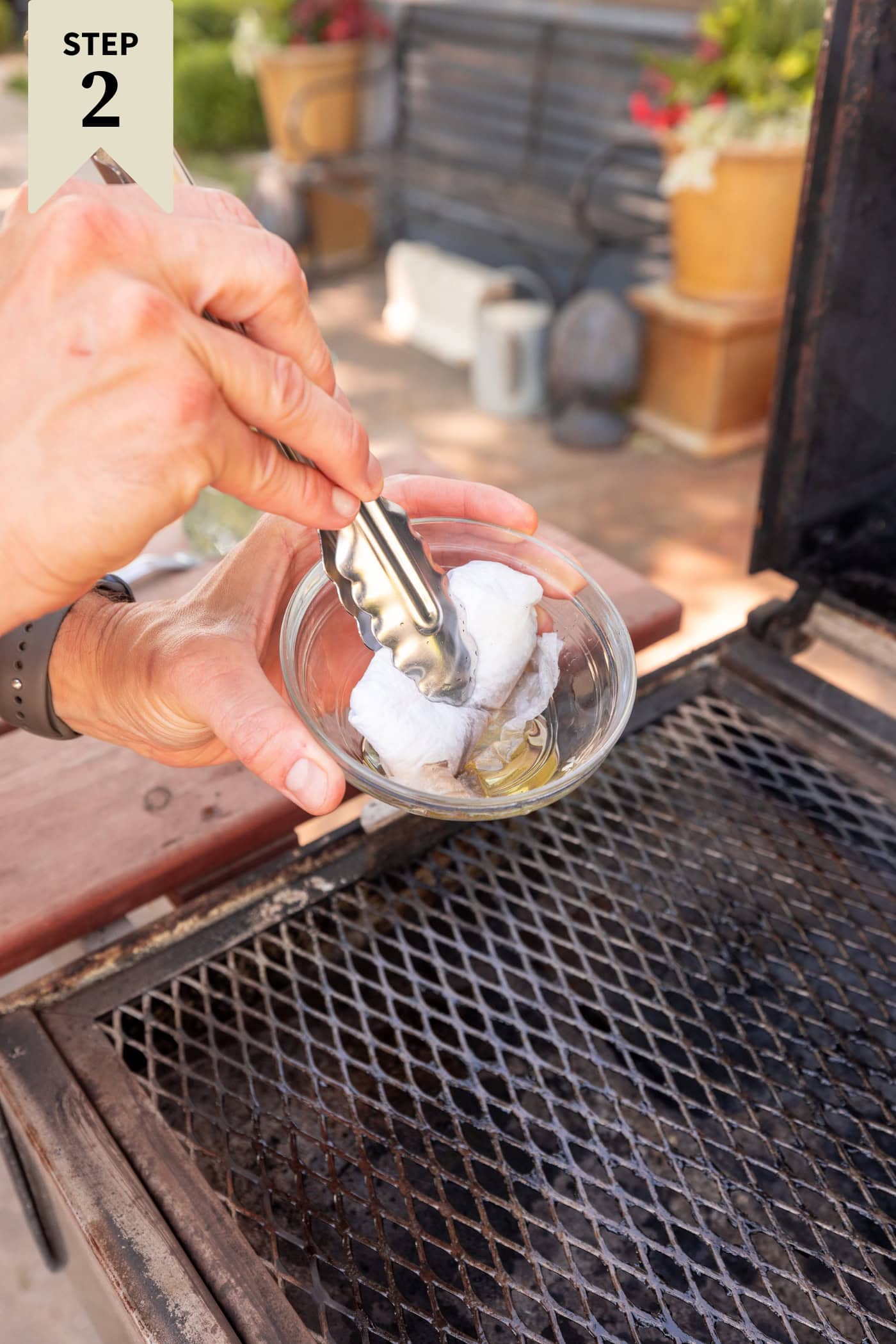 Hand using tongs to dip a paper towel into a small glass bowl filled with olive oil to brush on a grill.