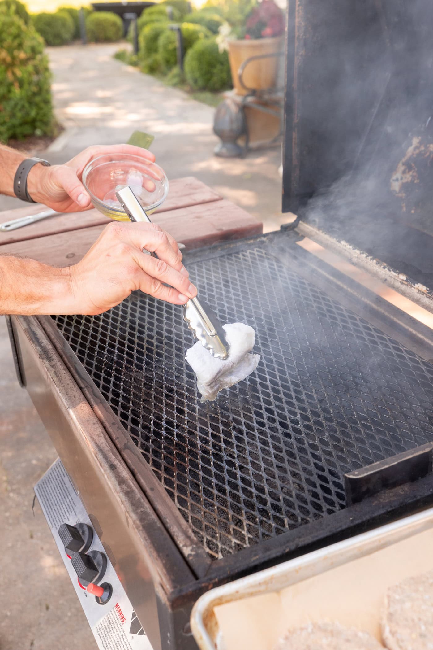Hand using tongs to brush an olive oil dipped paper towel on grates of a grill.