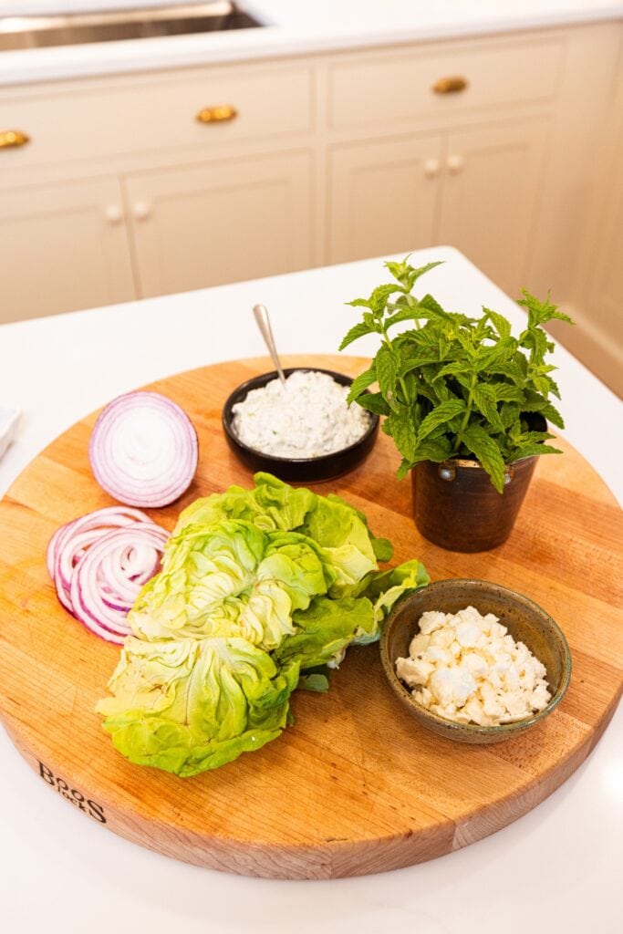 Wood cutting board filled with all ingredients to top a Greek turkey burger including lettuce, feta cheese, tzatziki sauce, and red onion.
