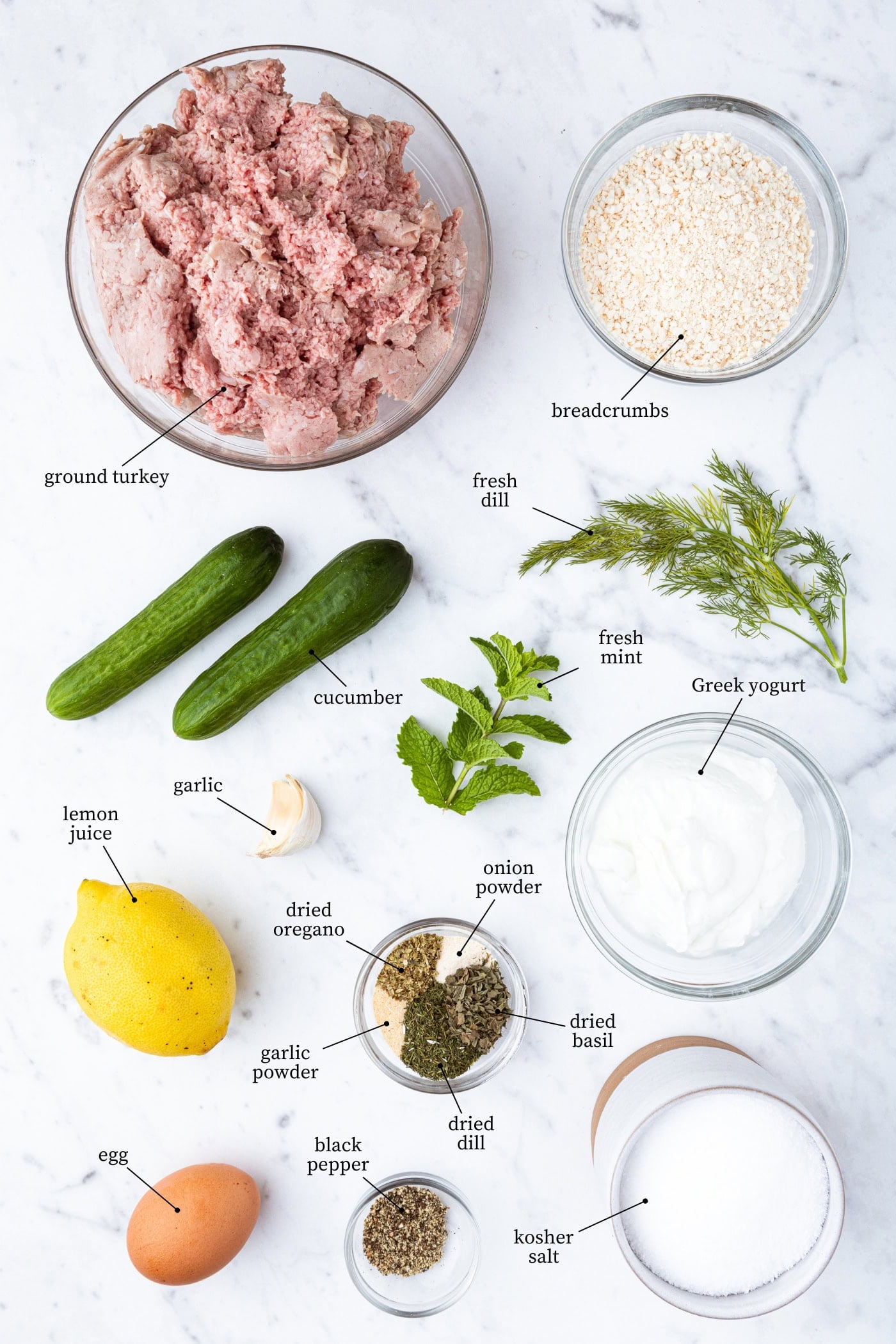 White marble surface filled with ingredients needed for Greek turkey burgers including raw turkey, breadcrumbs, cucumbers, lemon, herbs, spices, and more.