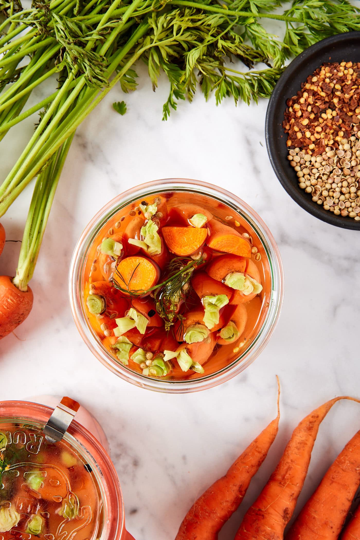 Glass jar filled with carrots in pickling brine with more jars and spices and carrots.