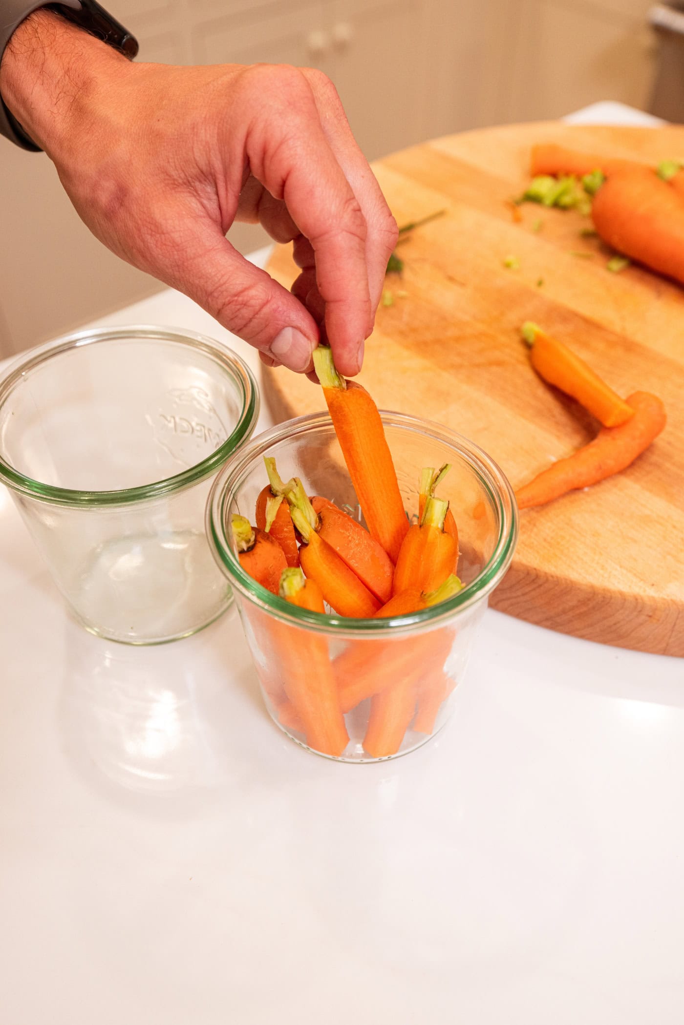 Hand placing raw carrots with a small amount of their tops still on in a glass jar.