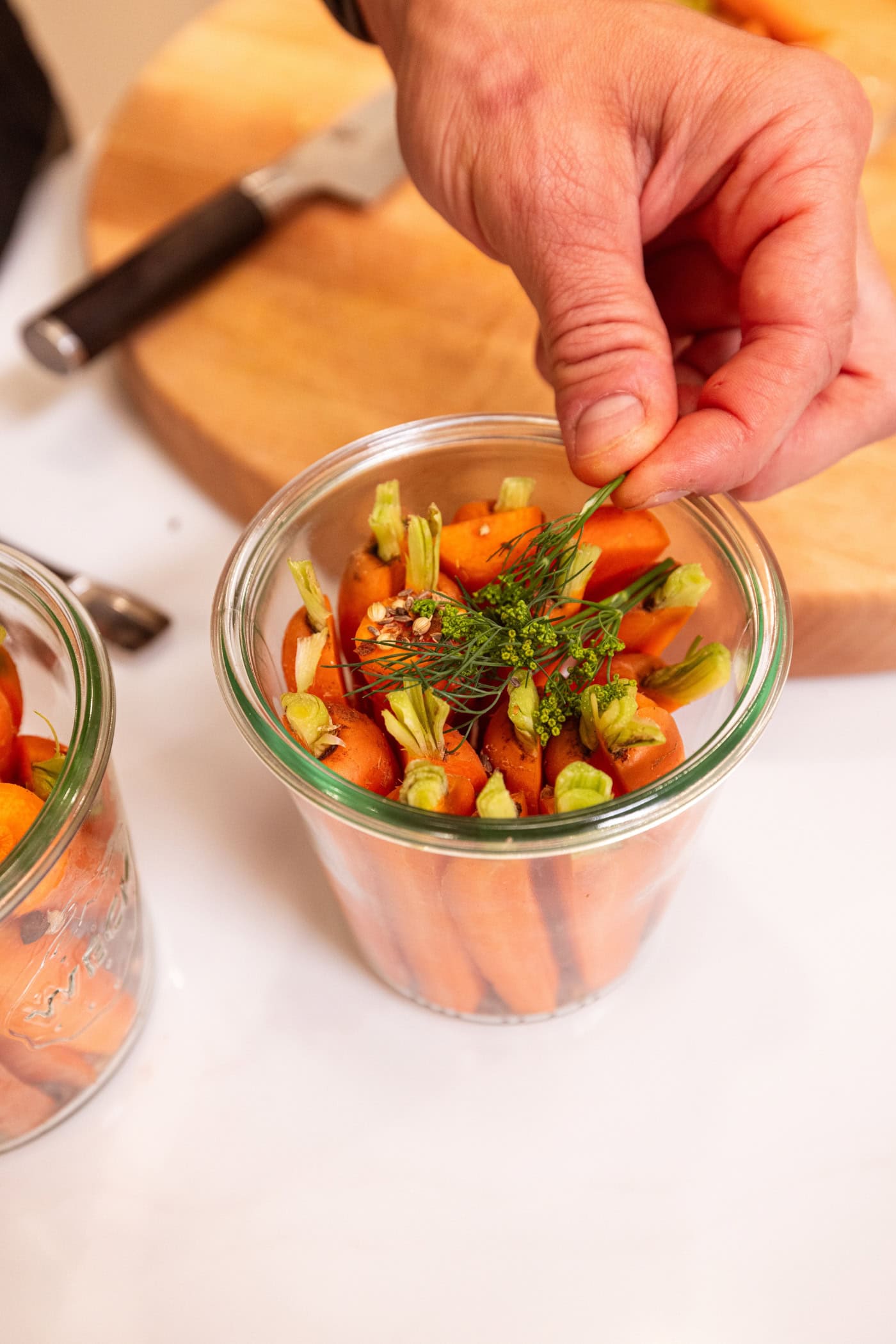 Hand holding sprig of fresh dill over top of glass jar filled with raw carrots.