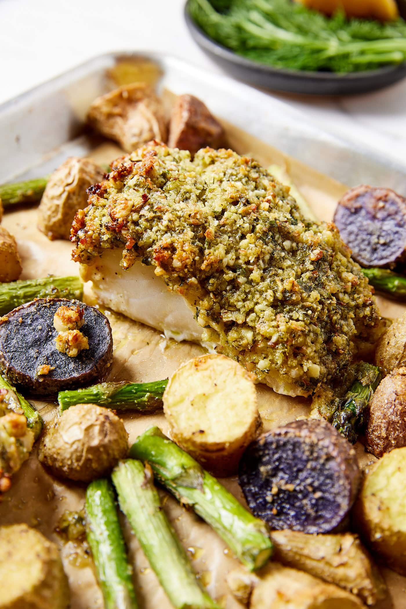 Piece of baked halibut sitting on sheet pan with roasted sliced potatoes and asparagus with herb crust on fish.