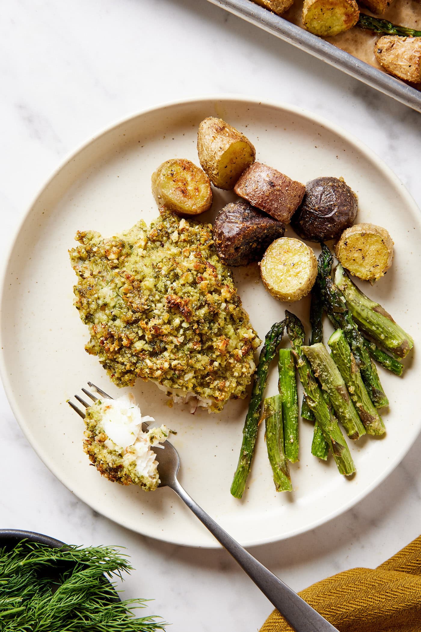 Plate with serving of baked halibut with roasted potatoes and asparagus on plate as well with bite on fork.