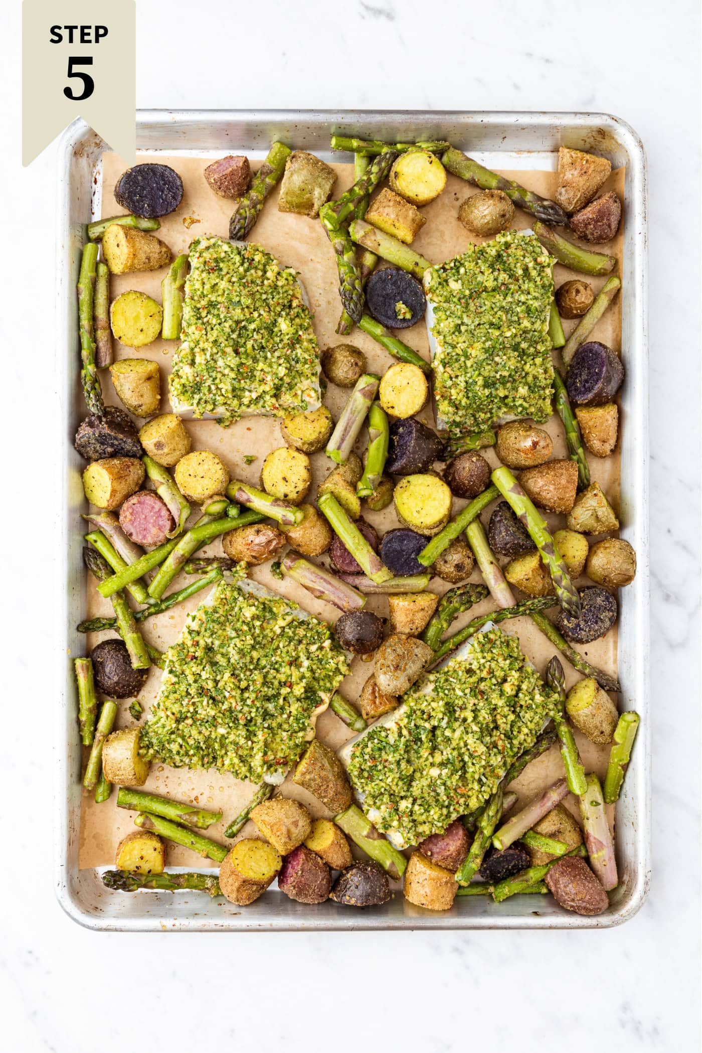 Sheet pan on white marble surface with halibut with herb crust and sliced potatoes and asparagus before being baked.