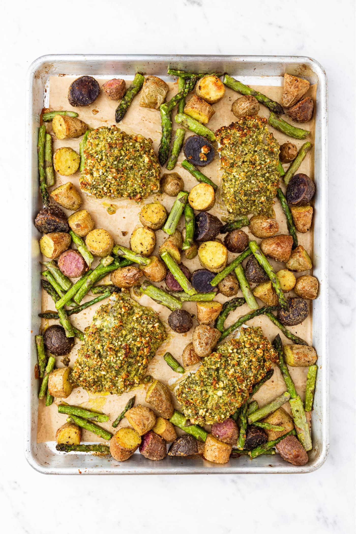 Sheet pan with baked halibut with herb crust on top with roasted asparagus and sliced potatoes.