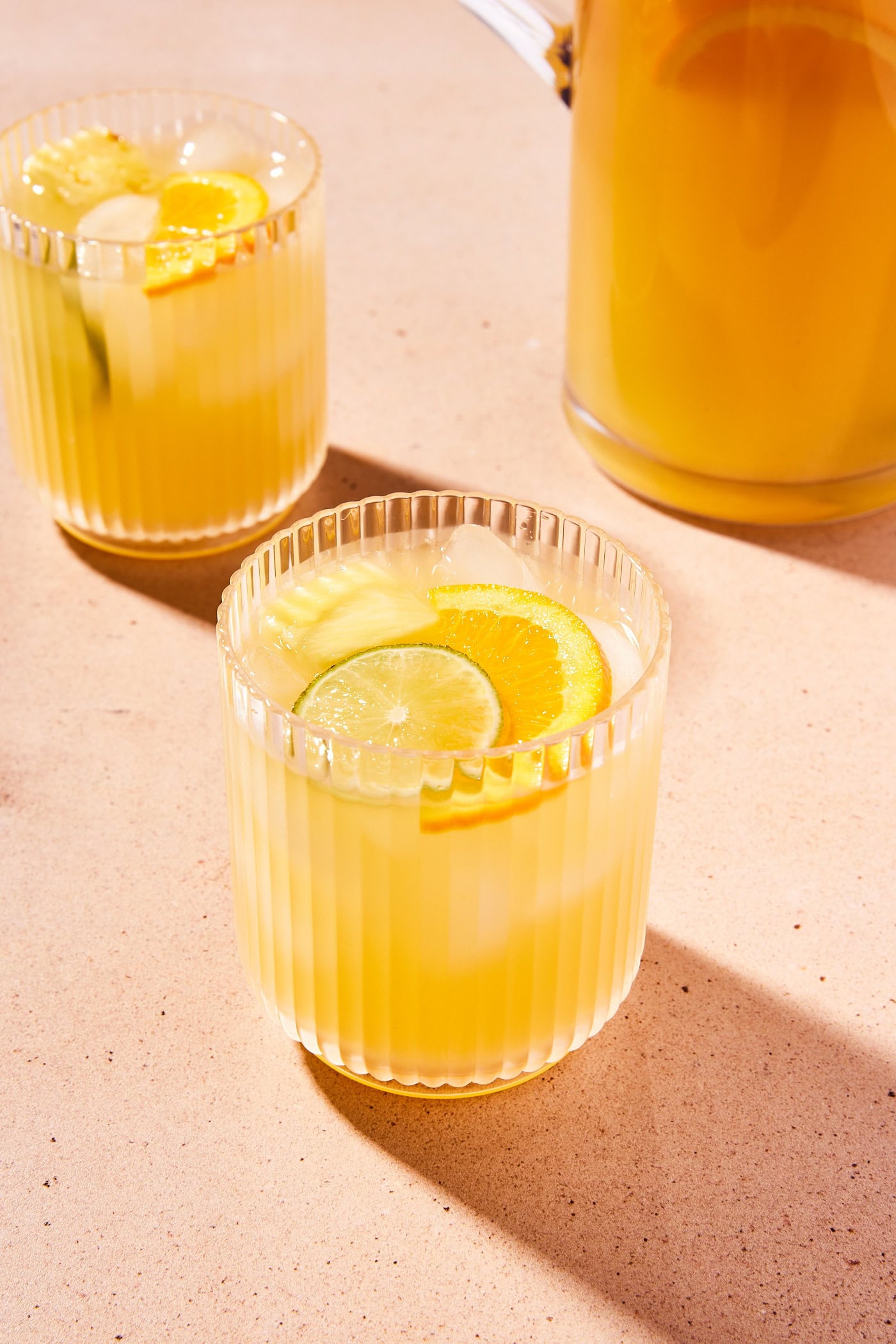 Two glasses of sangria with slices of orange and lime with pitcher filled with rest of liquid in background.