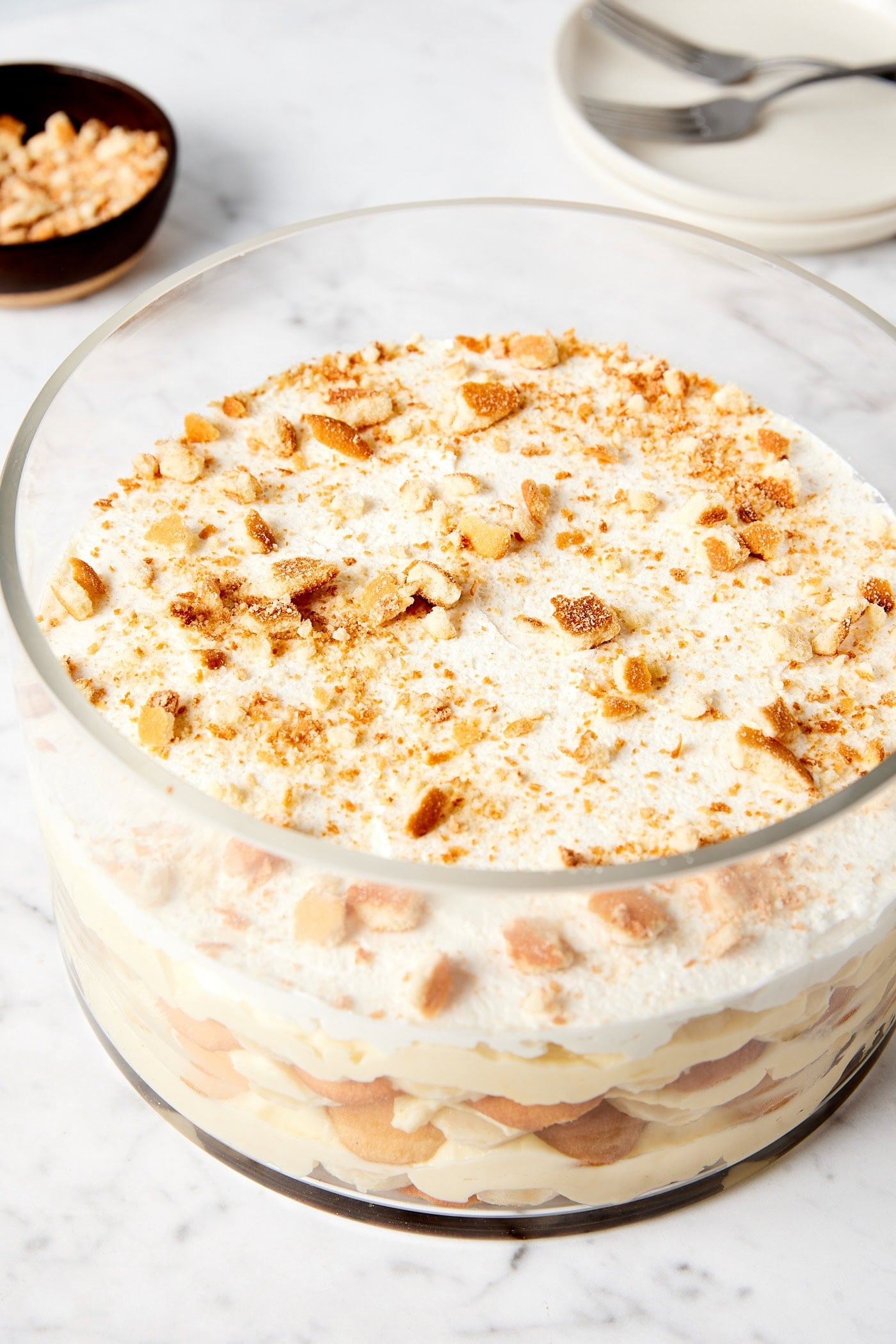 Glass dish filled with layers of banana pudding with plates and forks in the background all on white marble surface.