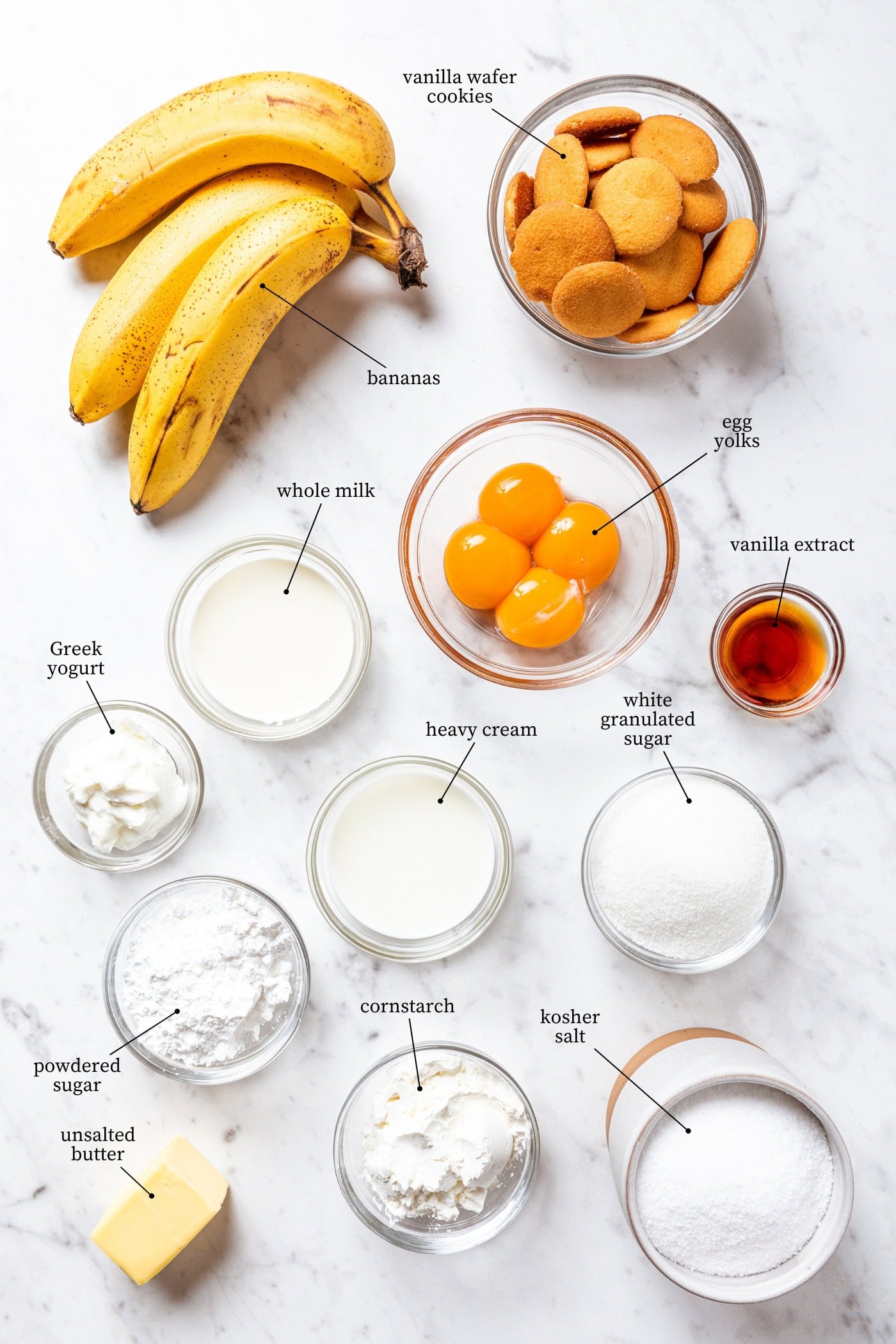White marble surface filled with all ingredients needed for a banana pudding recipe including bananas, vanilla wafers, egg yolks, sugar, and more.