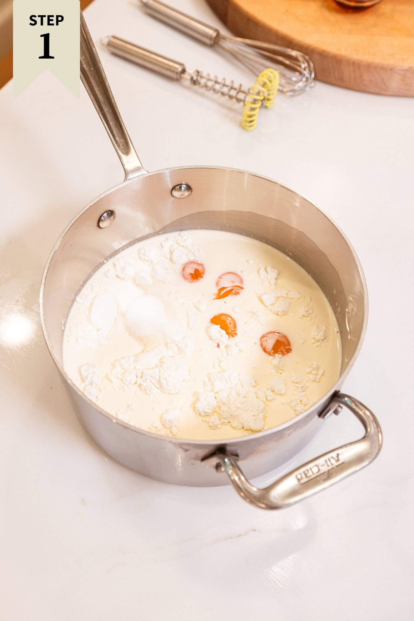 Metal saucepan on white surface with egg yolks, cream, milk, and sugar before being heated on the stovetop.