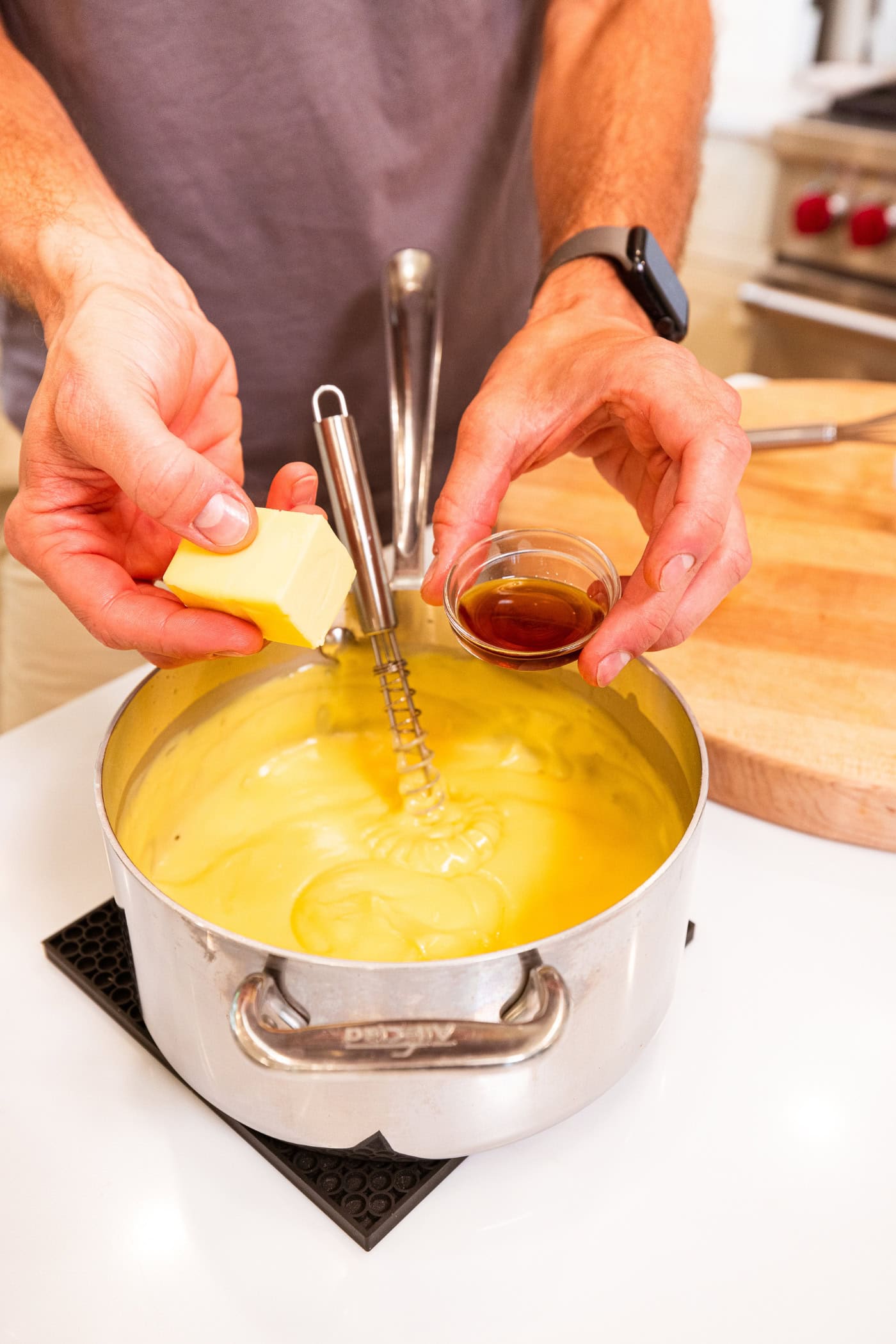 Hand holding butter and vanilla extract right before adding to vanilla pudding in a saucepan.