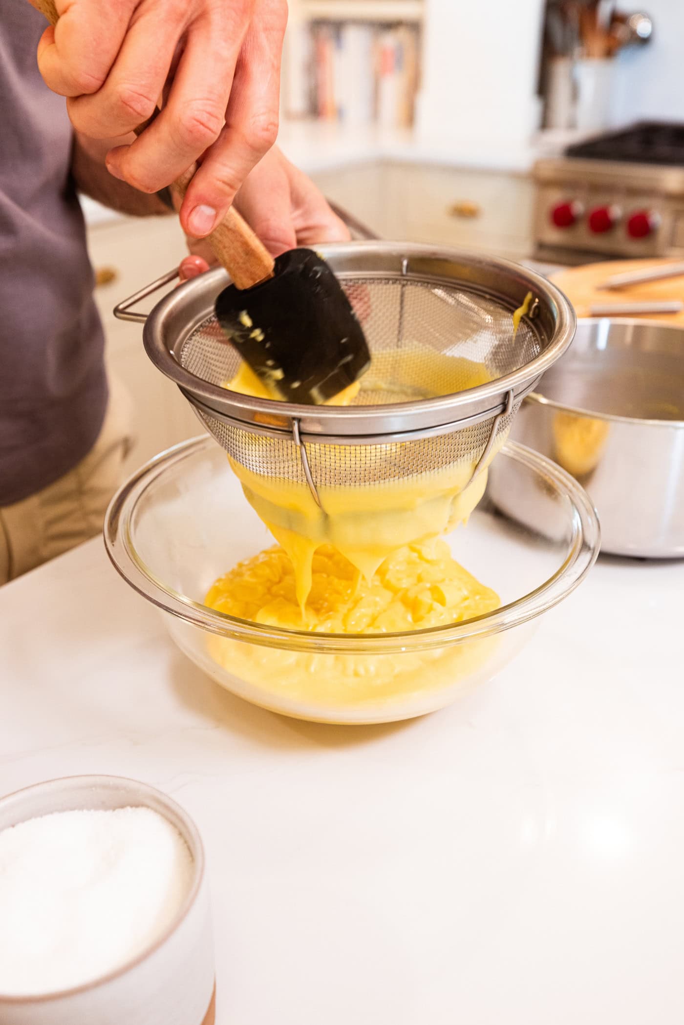 Hand using plastic spatula to push vanilla pudding through a strainer to ensure no lumps into a waiting glass bowl.