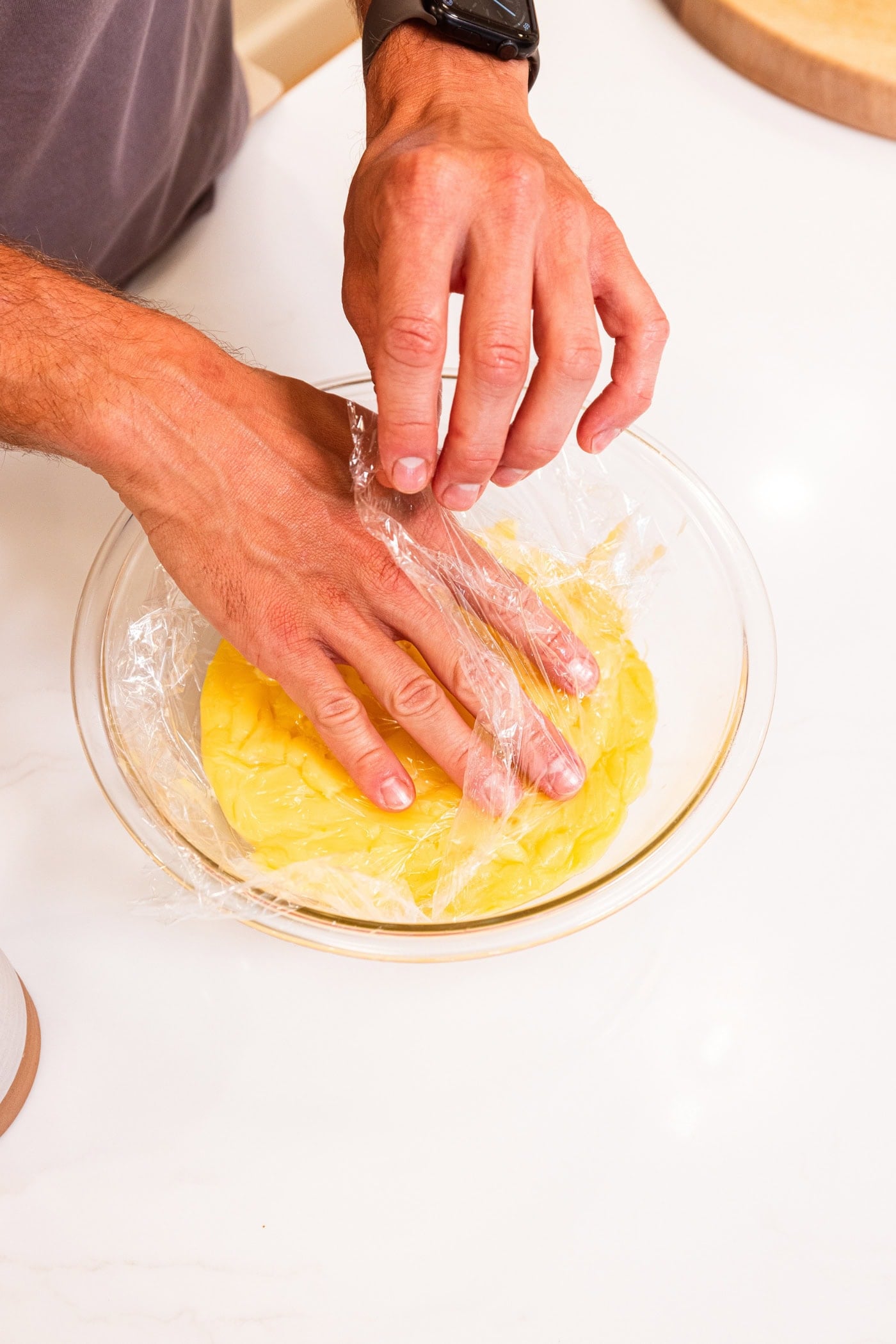 Hand placing plastic wrap on top of vanilla pudding to prevent a skin from forming.
