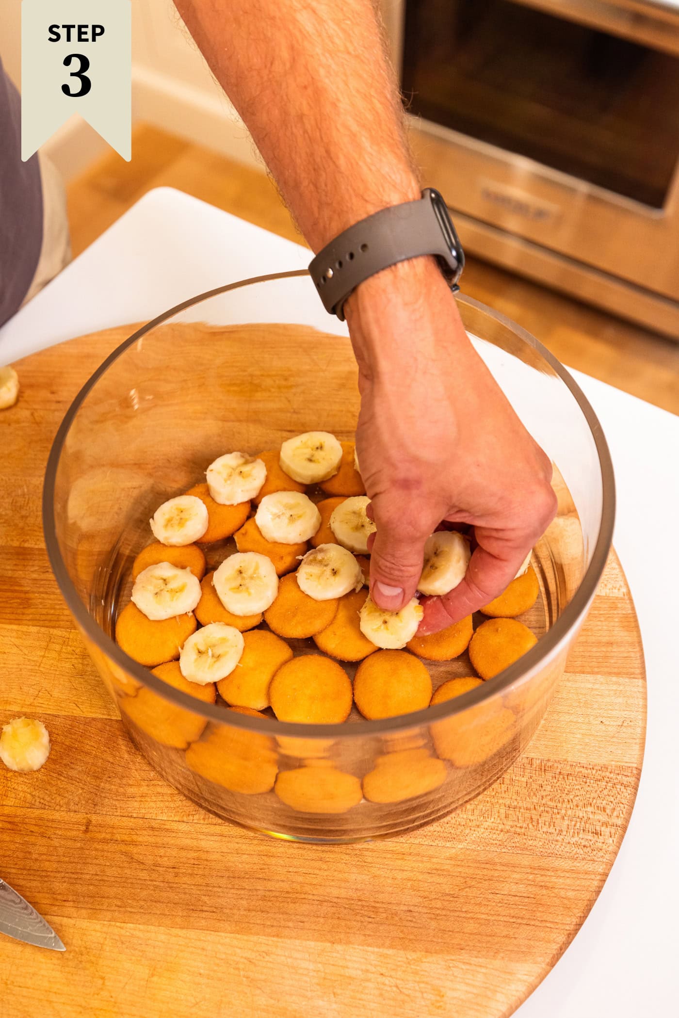 Hand placing slices of banana on top of vanilla wafer cookies in a glass trifle pan.