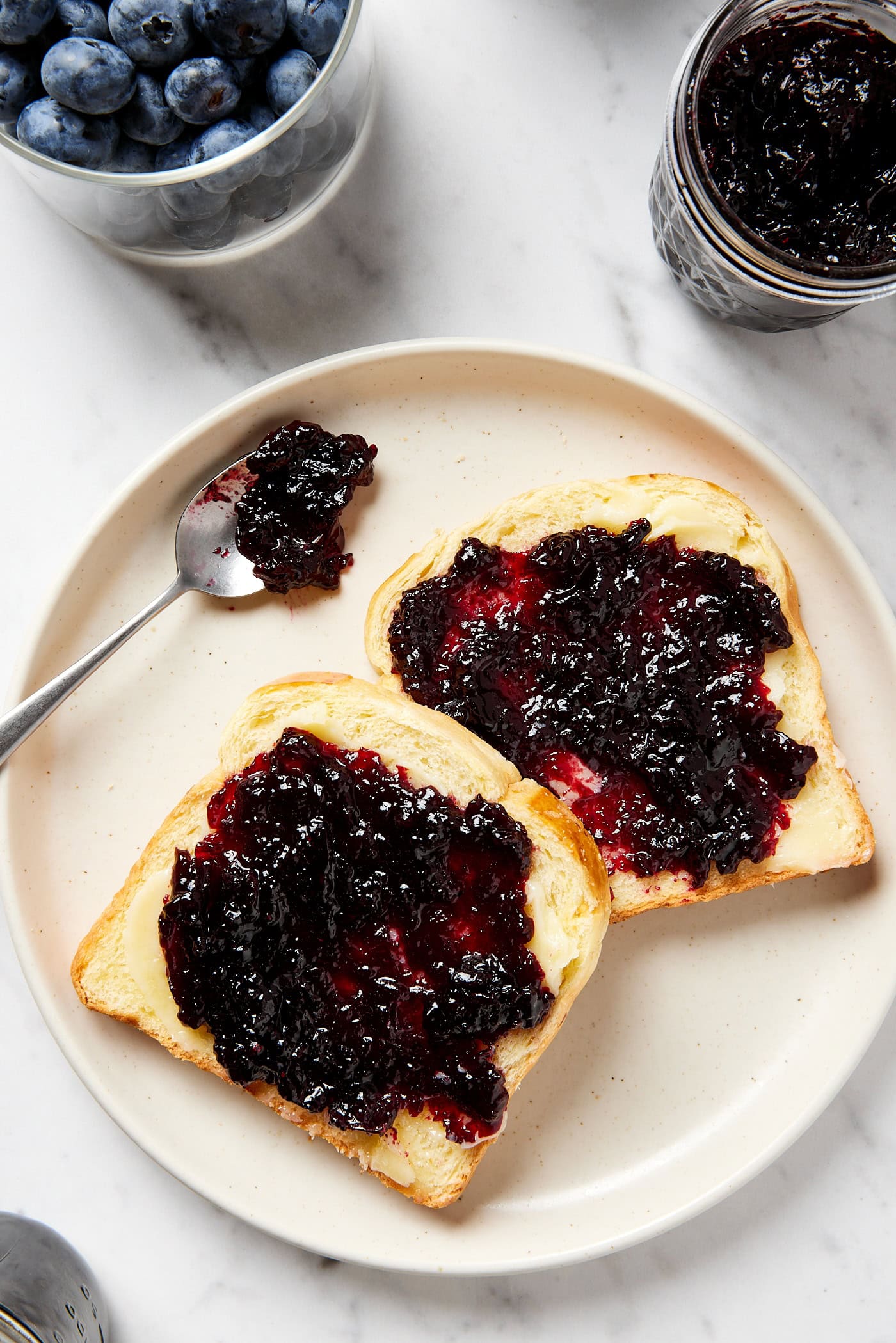 White plate with two slices of buttered bread with spoon resting on plate after spreading jam on top.