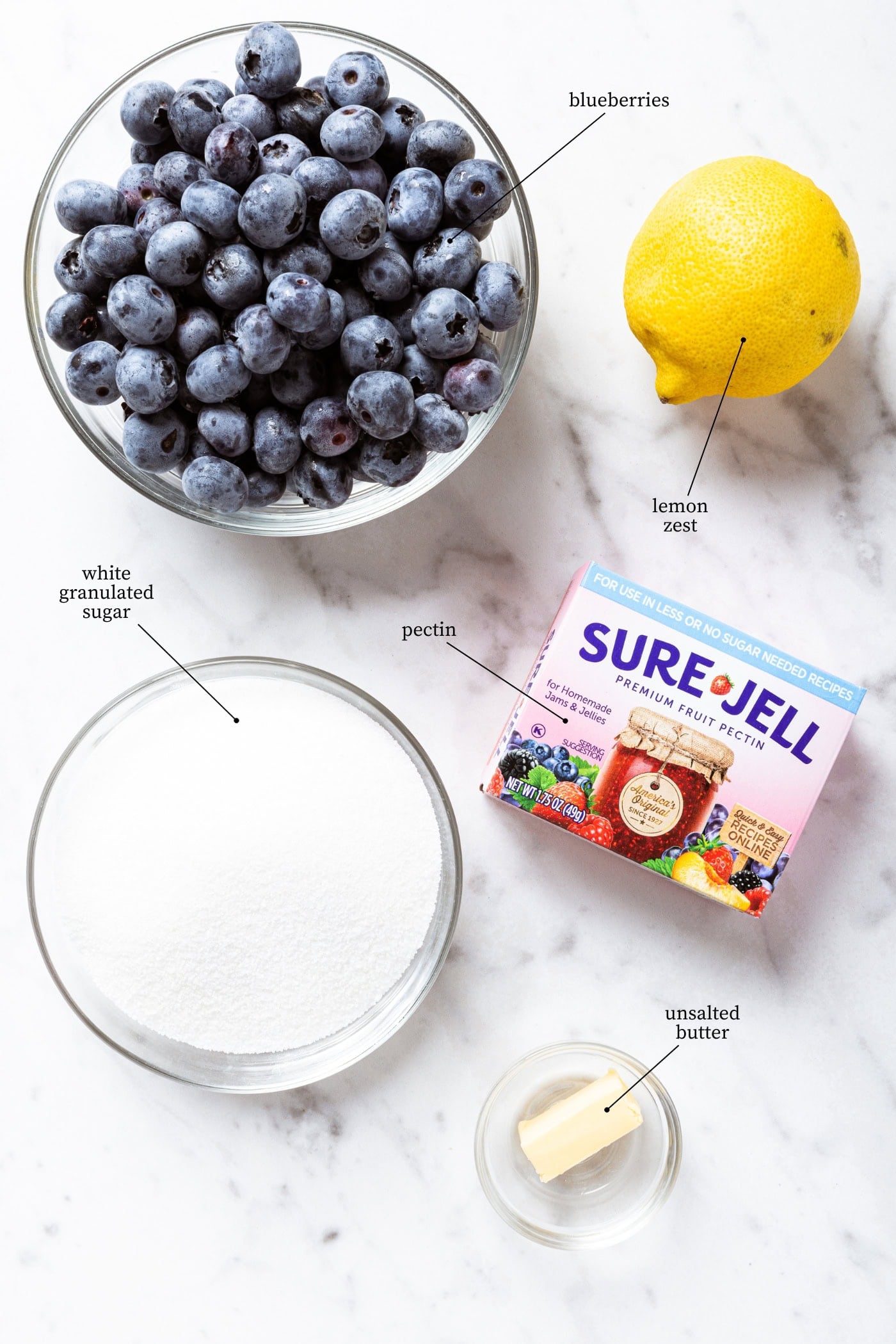 White marble surface filled with all ingredients needed to make blueberry jam including blueberries, lemon, sugar, pectin, and butter.