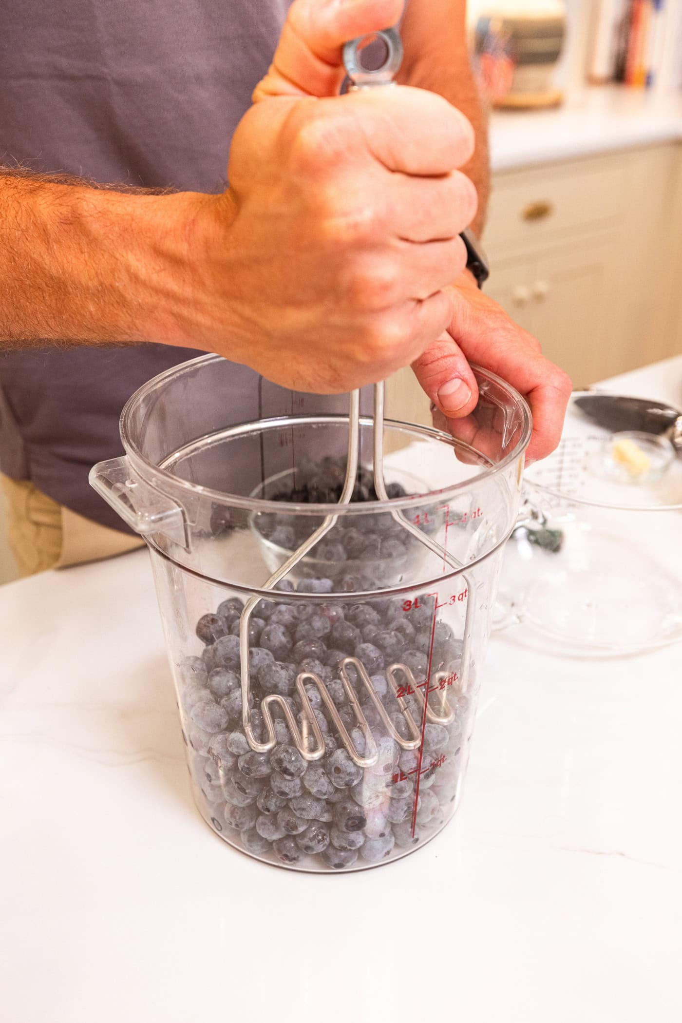 Hand using masher to mash blueberries in a large plastic container.