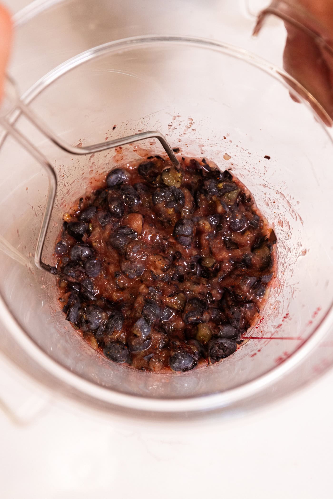 Mashed blueberries in large plastic container with masher resting in container.