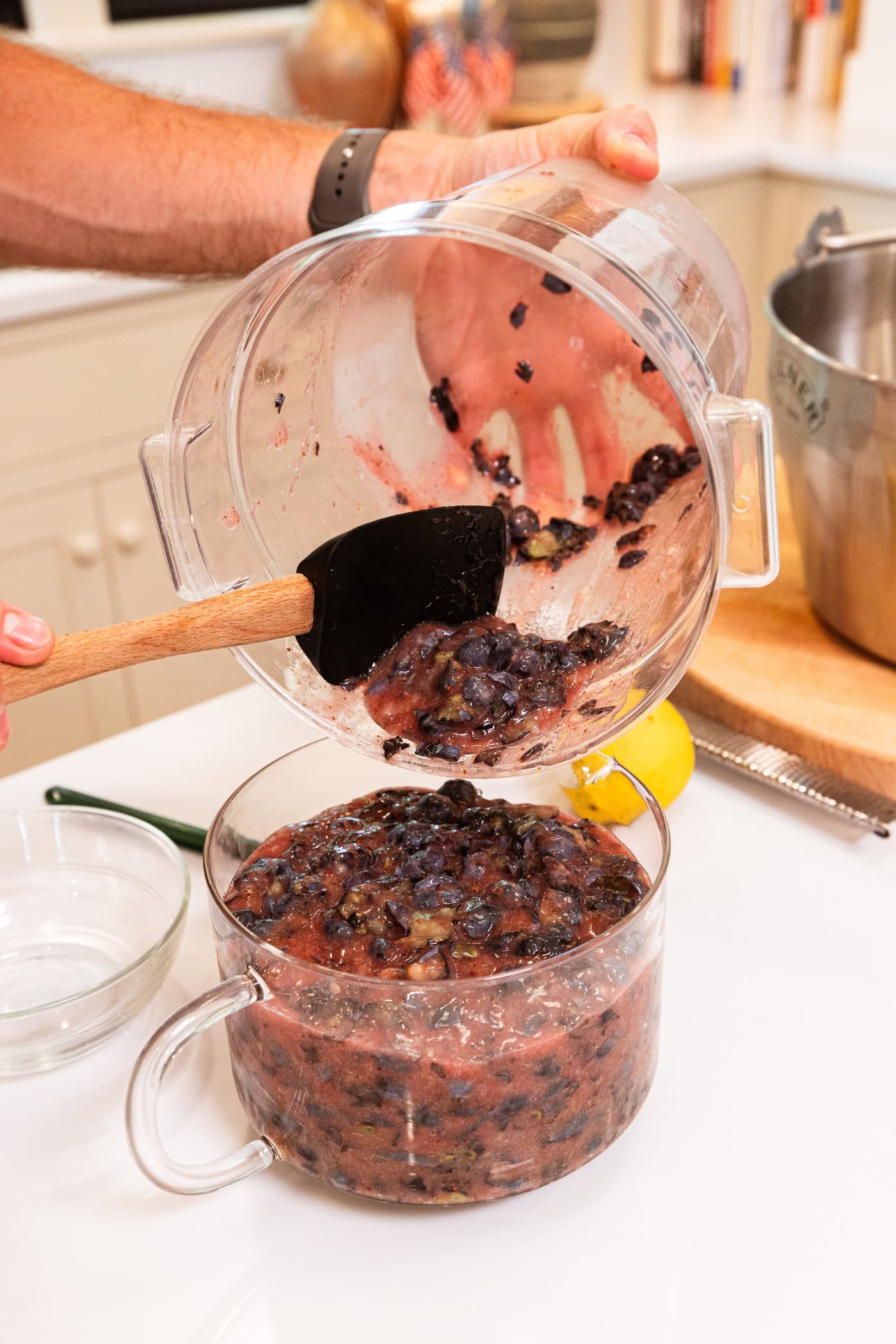Hand holding container measuring mashed blueberries into a glass measuring cup.