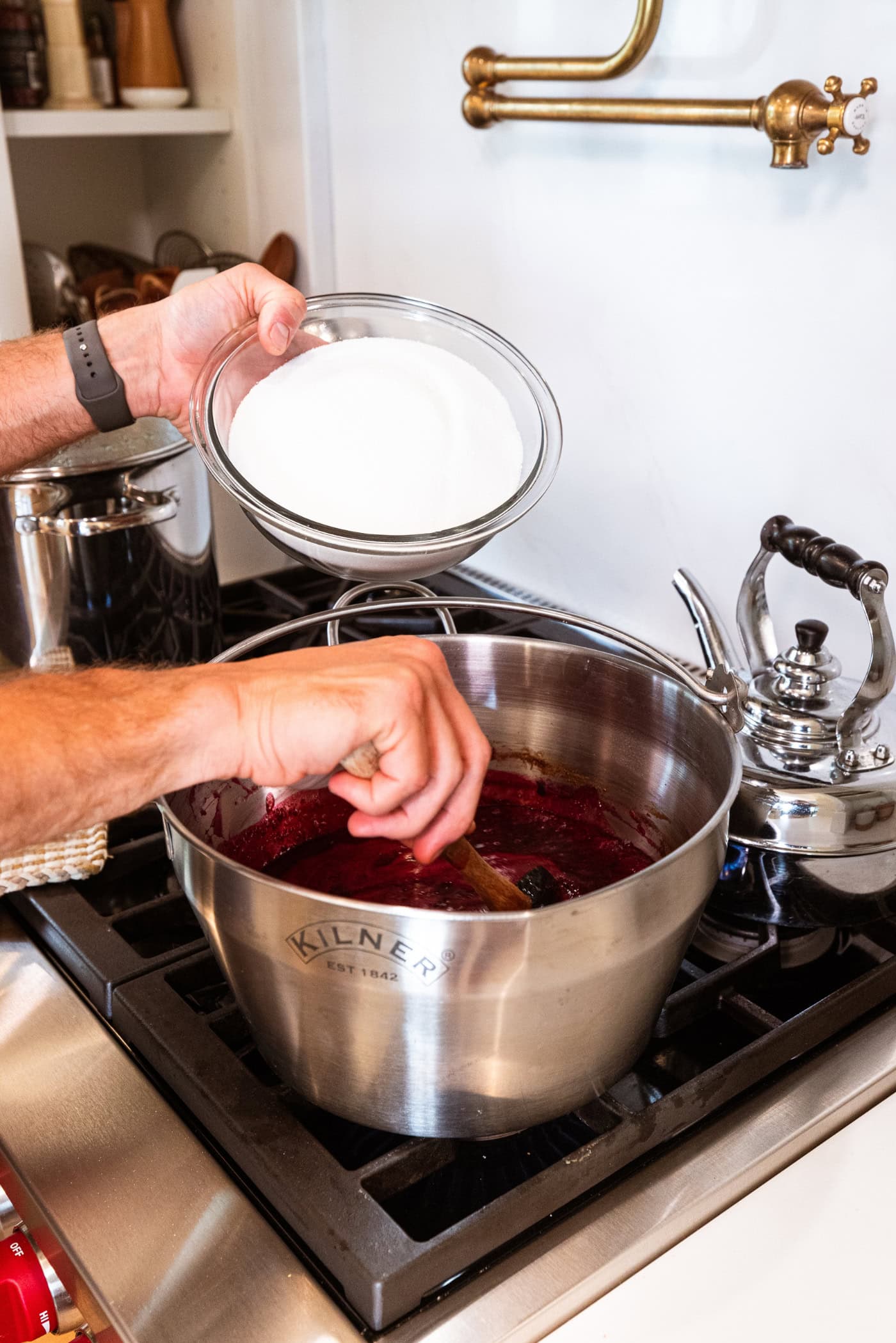 Hand holding glass bowl filled with sugar about to pour it into a large metal container on the stovetop.