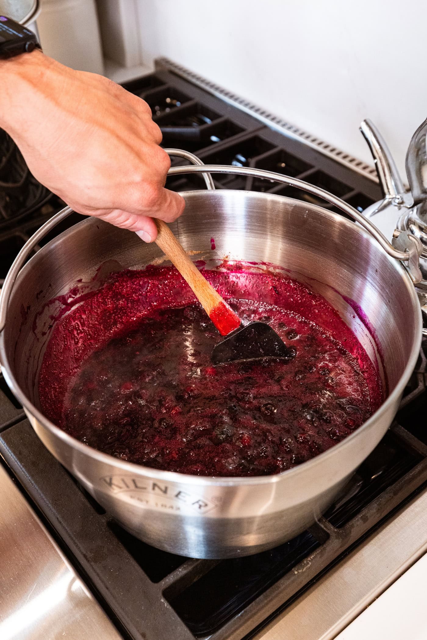 Hand using spatula to mix together sugar with blueberries to form a jam.