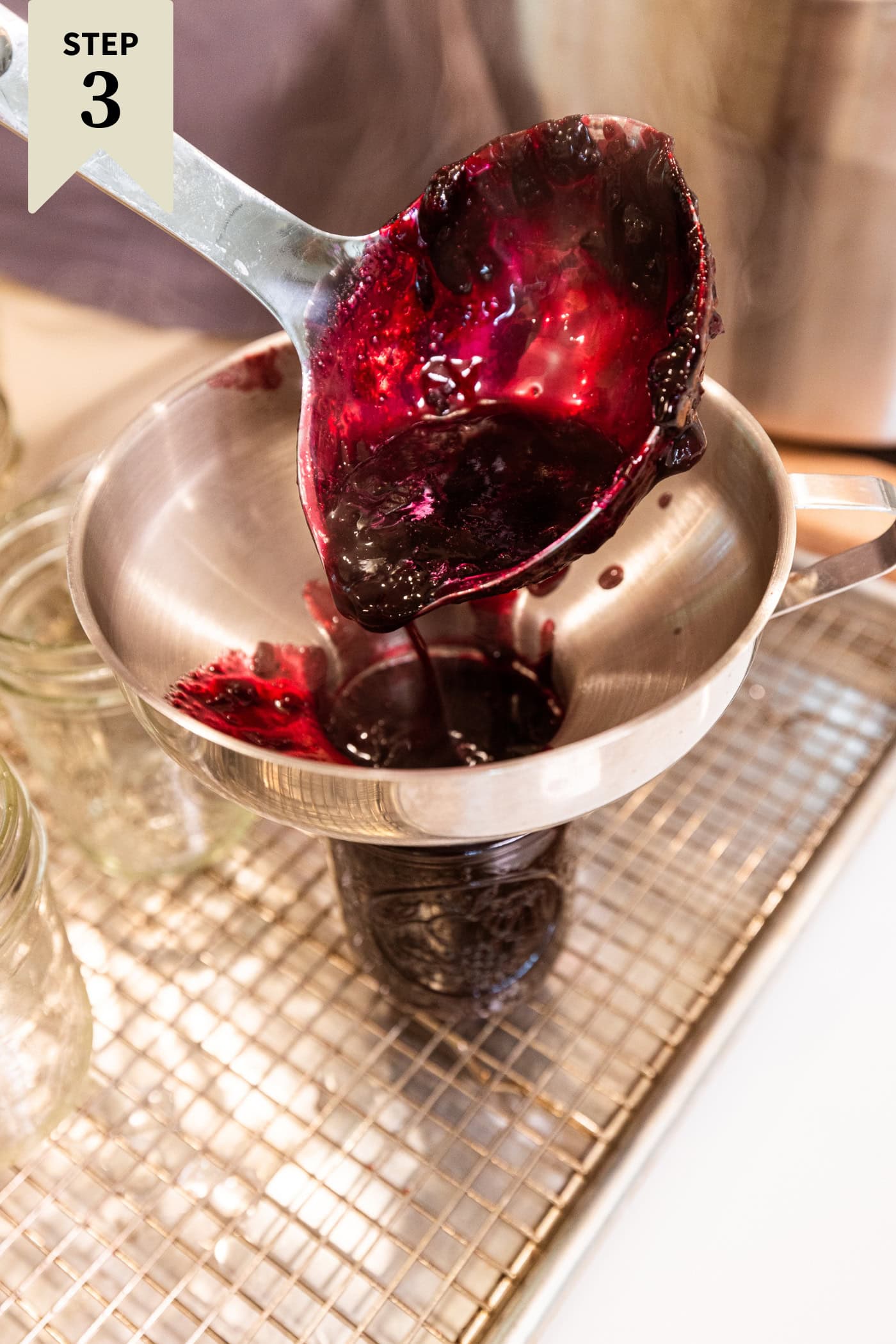 Ladle pouring cooked blueberry jam into a jam jar through a funnel.