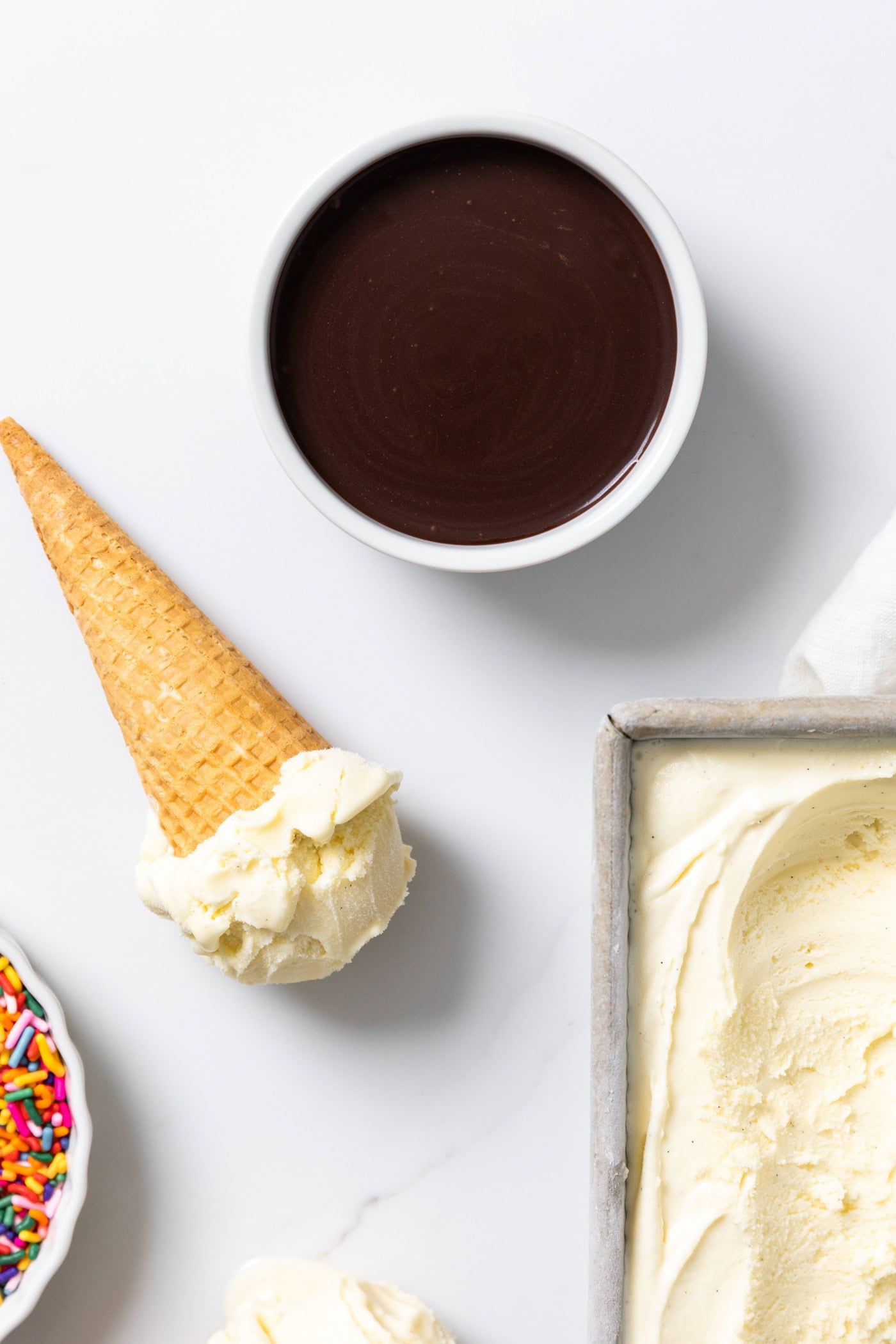 Bowl of chocolate hot fudge sauce sitting on white surface beside an ice cream cone.