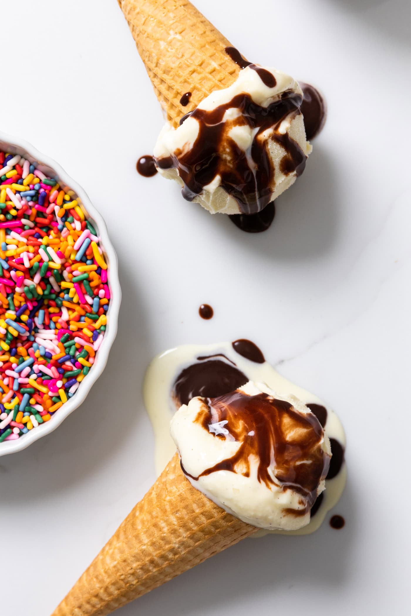 Two vanilla ice cream cones with hot fudge dripping off the cones onto white surface with sprinkles to the side.