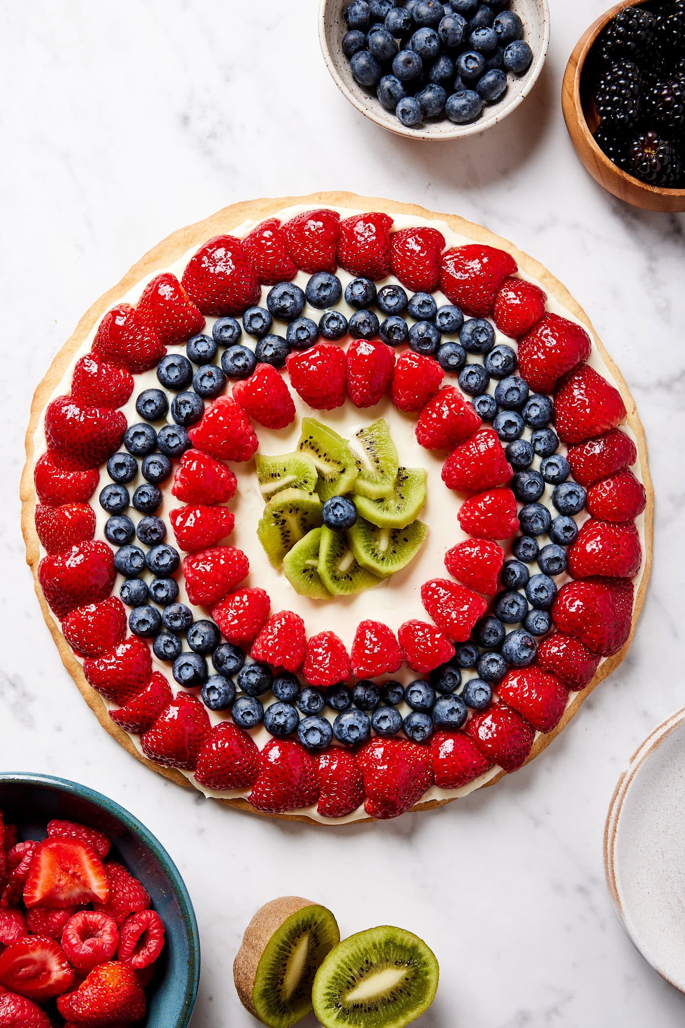 Round fruit pizza decorated with lots of fruit on white marble surface with more fruit yet to be added.