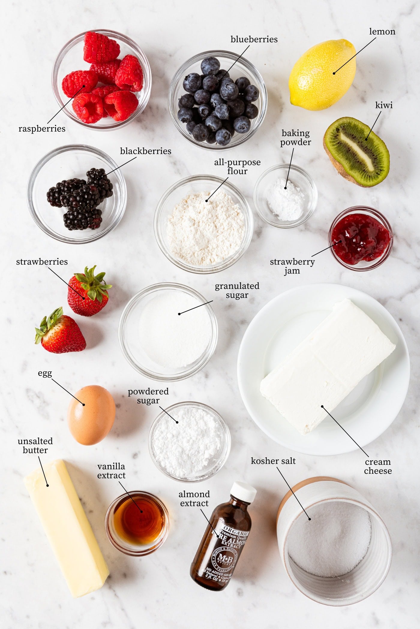 White marble surface with all ingredients needed to make fruit pizza including fruit, cream cheese, sugar, butter, egg, salt, and more.