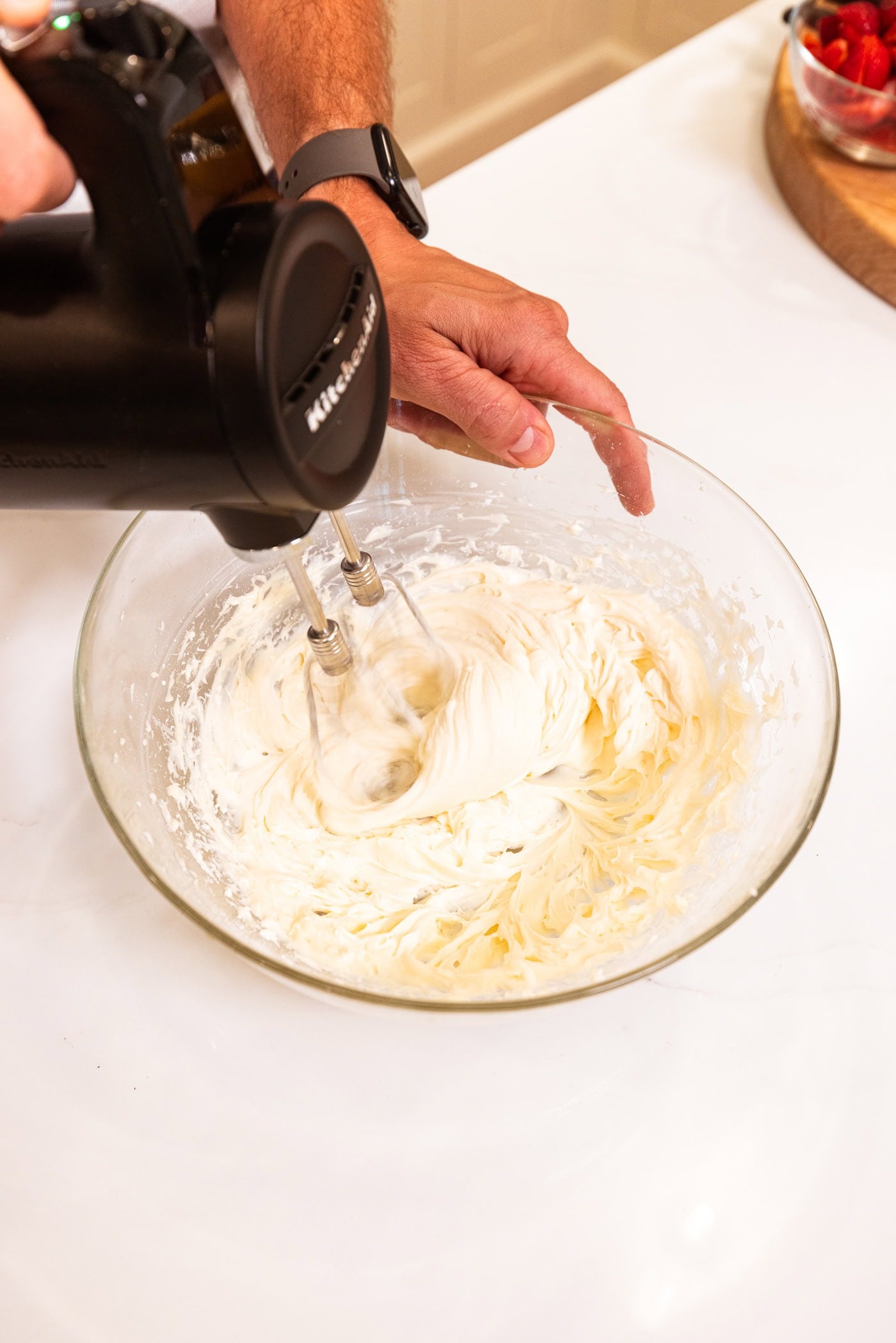 Hand mixer mixing together cream cheese frosting in a glass bowl on a white surface.