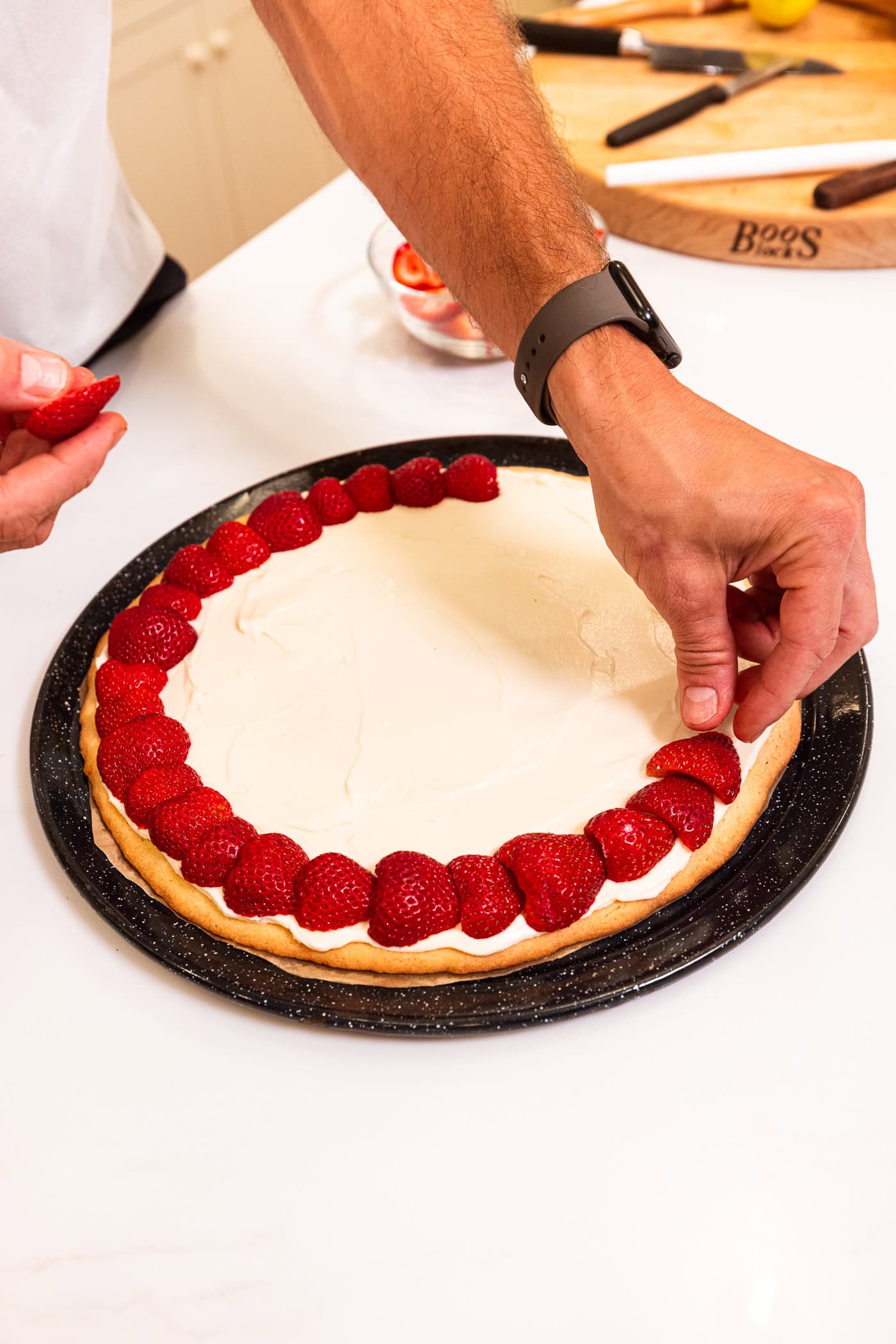 Hand placing strawberries on top of a cream cheese frosting on a sugar cookie crust.